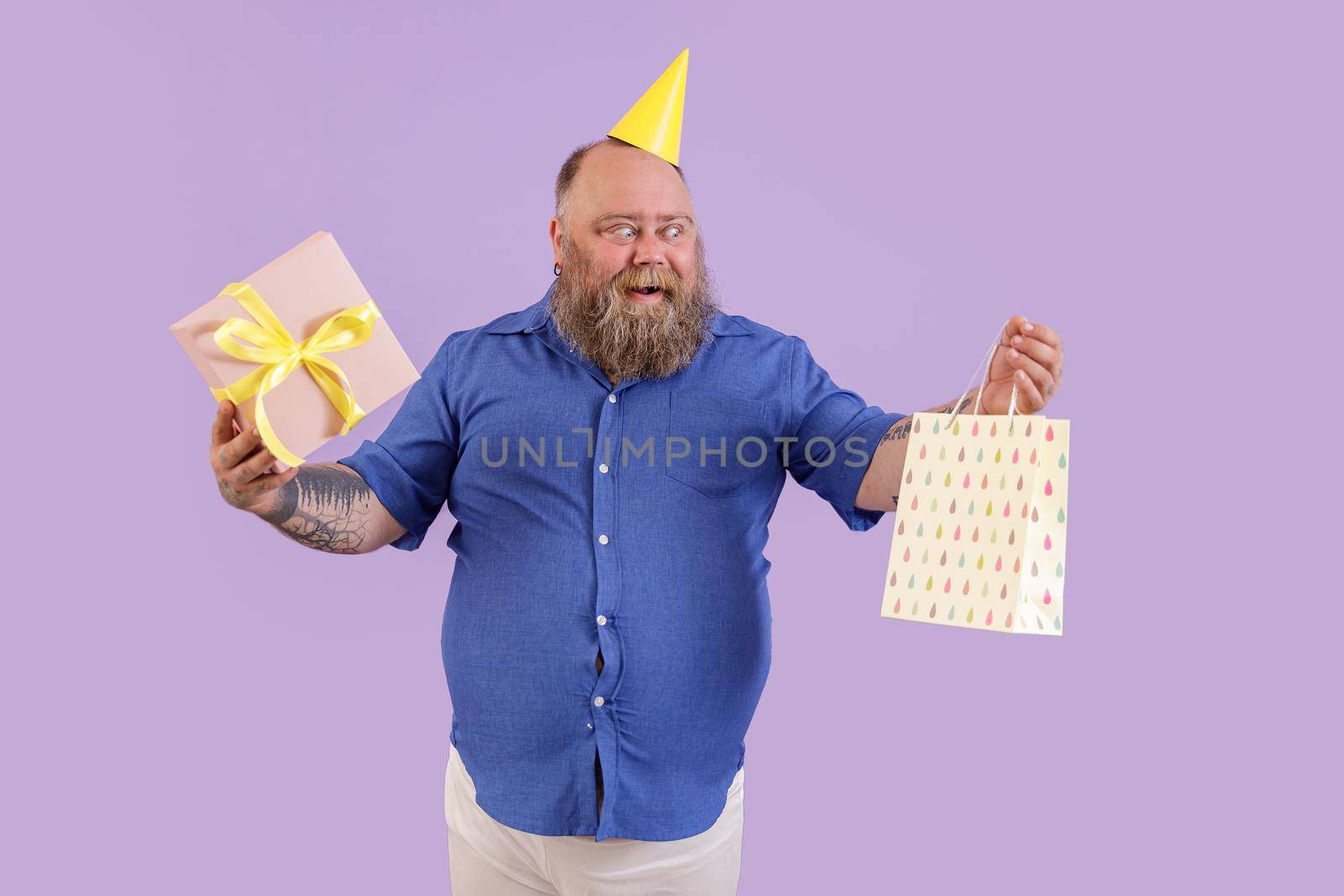 Excited mature bearded fat man in yellow party hat holds gift box and paper bag standing on purple background in studio
