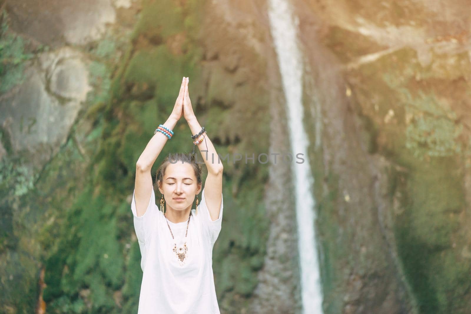 Young woman relaxing and practicing yoga with closed eyes near the waterfall in summer outdoor.