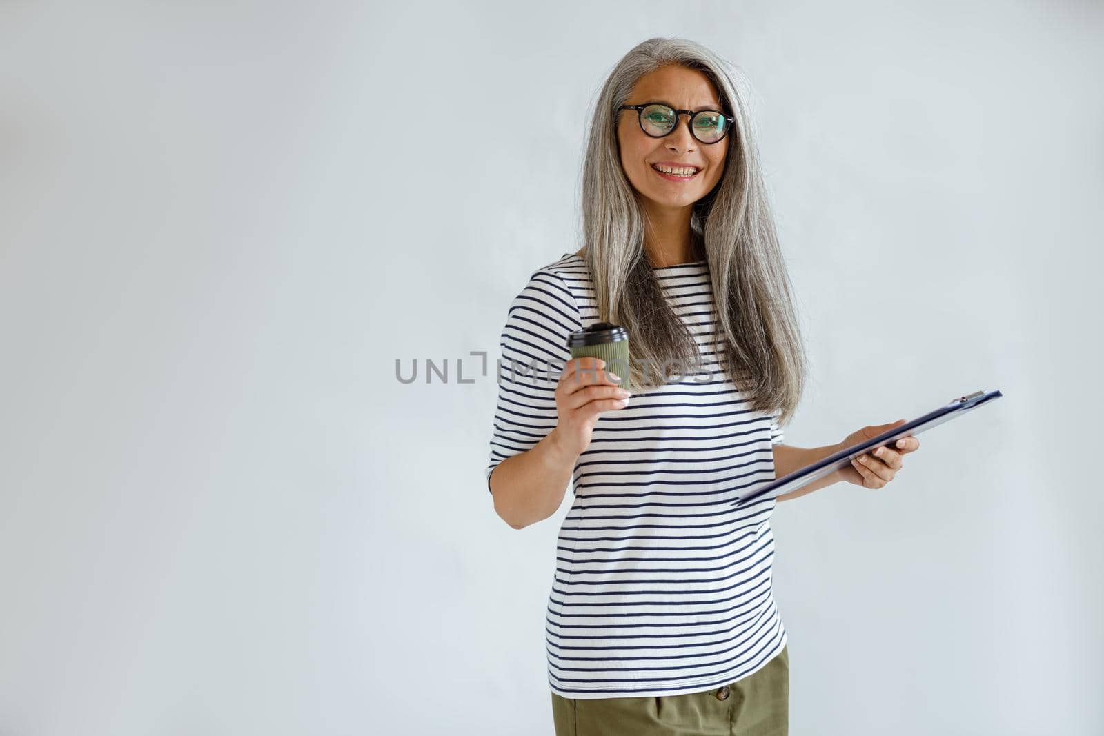 Cheerful mature Asian lady with natural silver hair holds clipboard and cup standing on light grey background in studio, space for text