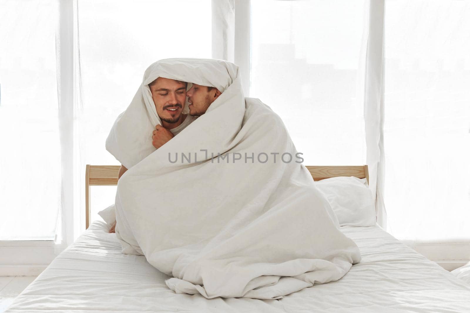 Loving gay men wrapping in cozy blanket and caressing on bed at home