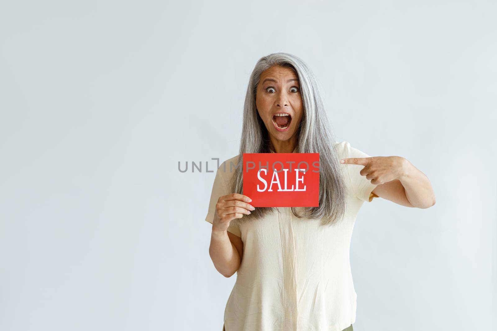 Excited mature Asian woman with long silver hair points onto red Sale sign posing on light background in studio, space for text