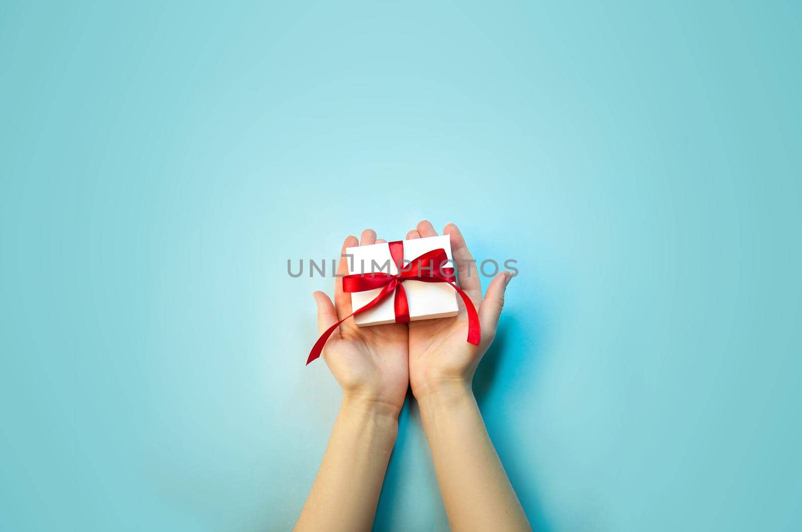 Christmas Holiday Composition. New Year Gift in White Box with Red Ribbon in Female Hands on Light Blue Background Flat Lay Top View with Copy Space For Your Text