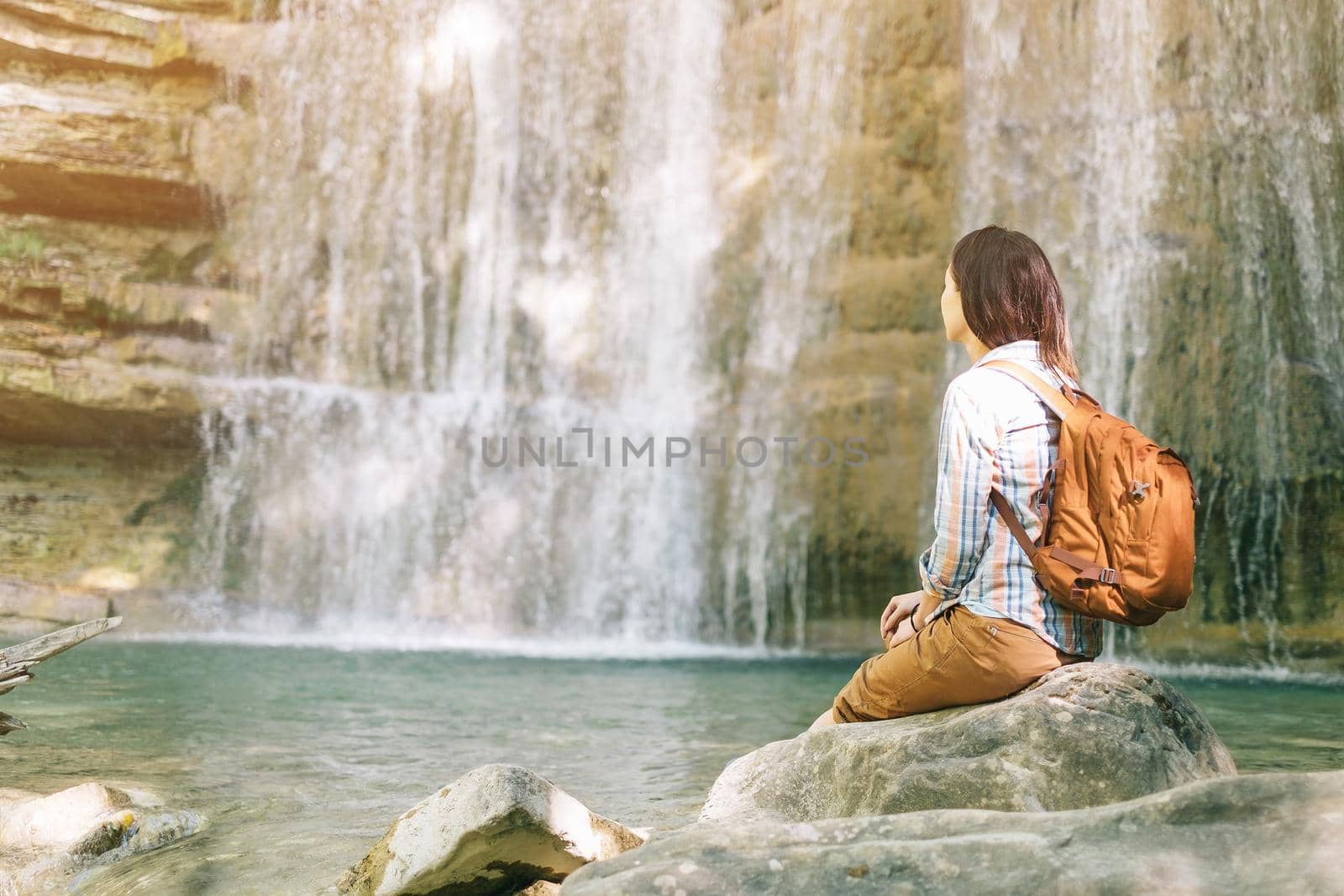 Backpacker young woman resting in front of waterfall on sunny day.