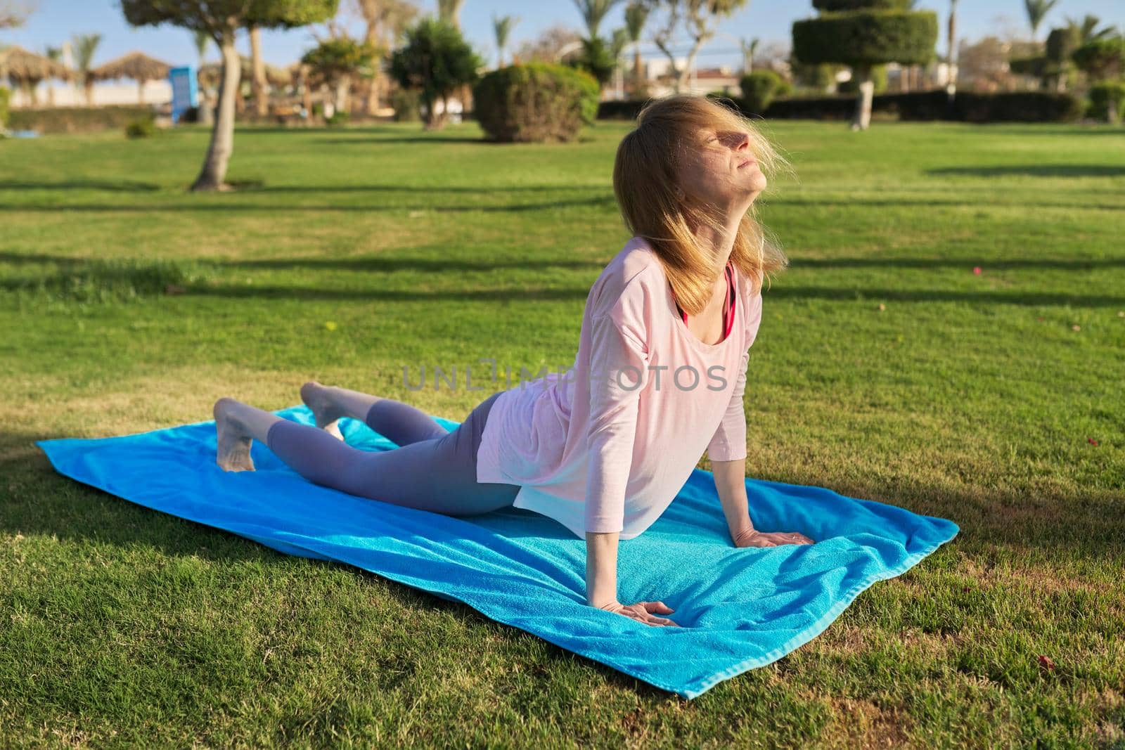 Middle-aged active woman doing sports exercises on a rug on the grass, in the morning. by VH-studio