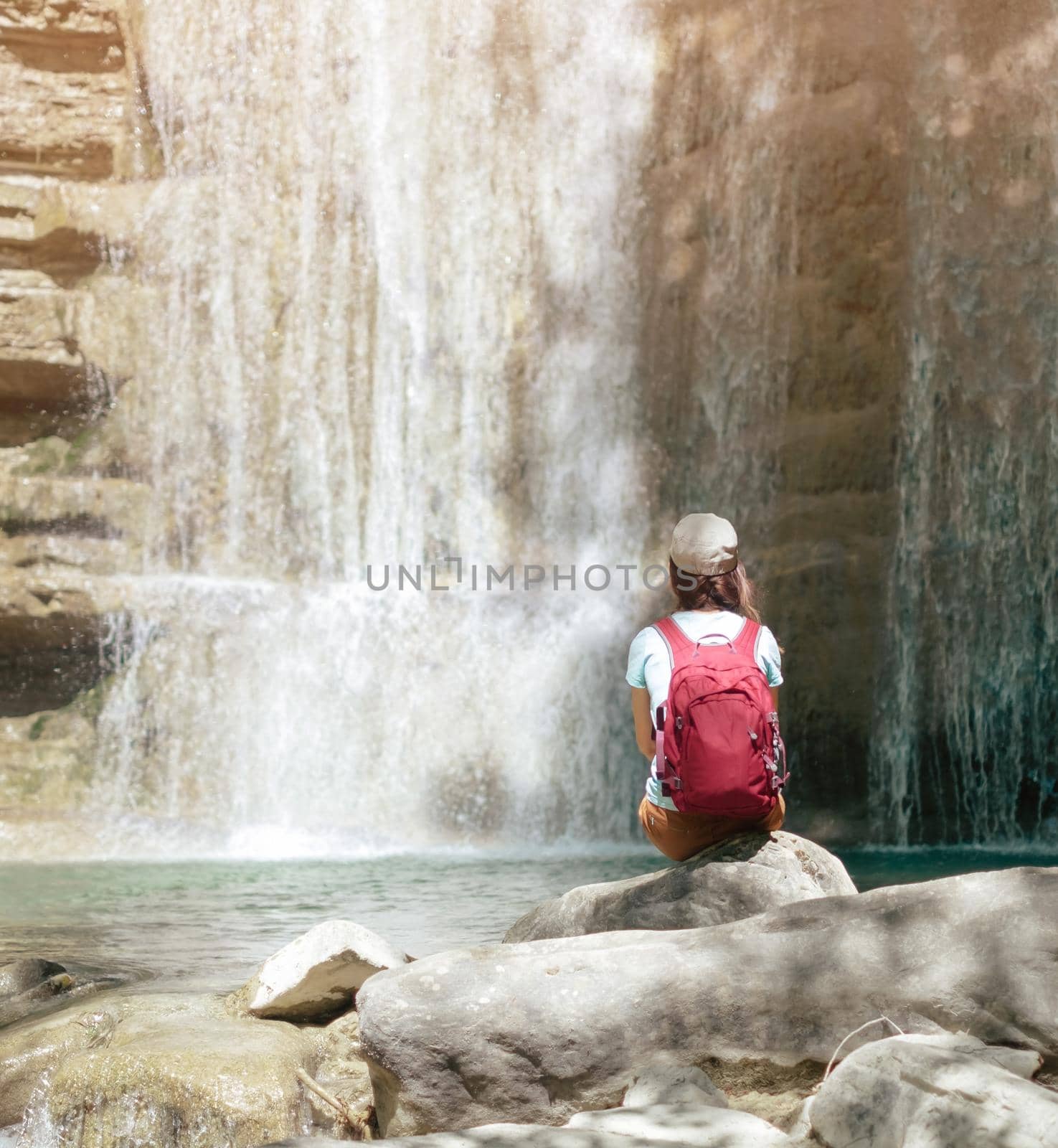 Female backpacker explorer sitting on stone and enjoying view of waterfall, rear view.