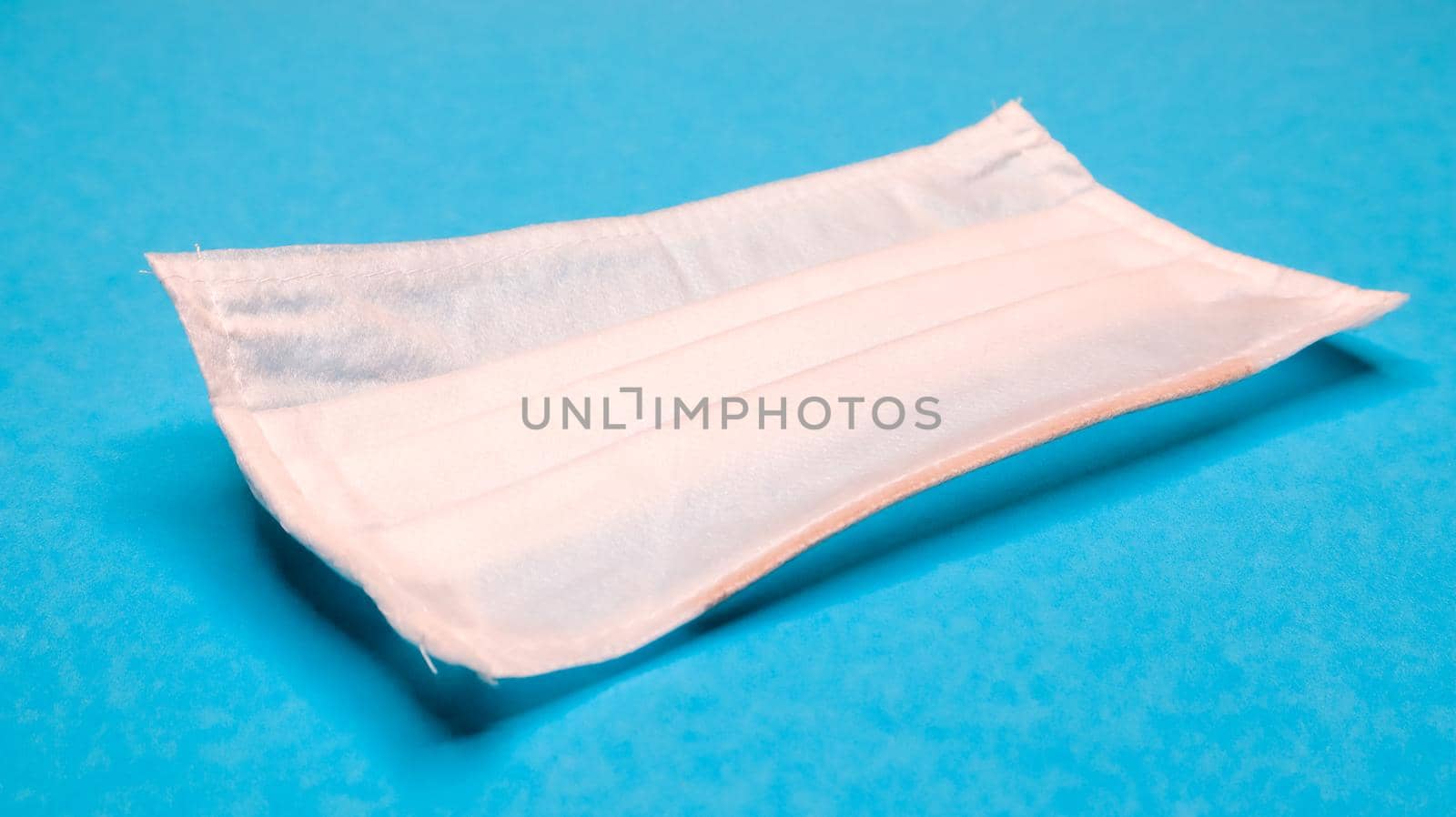 One 3-ply disposable surgical face mask with rubber grips covering mouth and nose against blue background. Protection from bacteria, healthcare and medicine concept. COVID-19. A dangerous virus