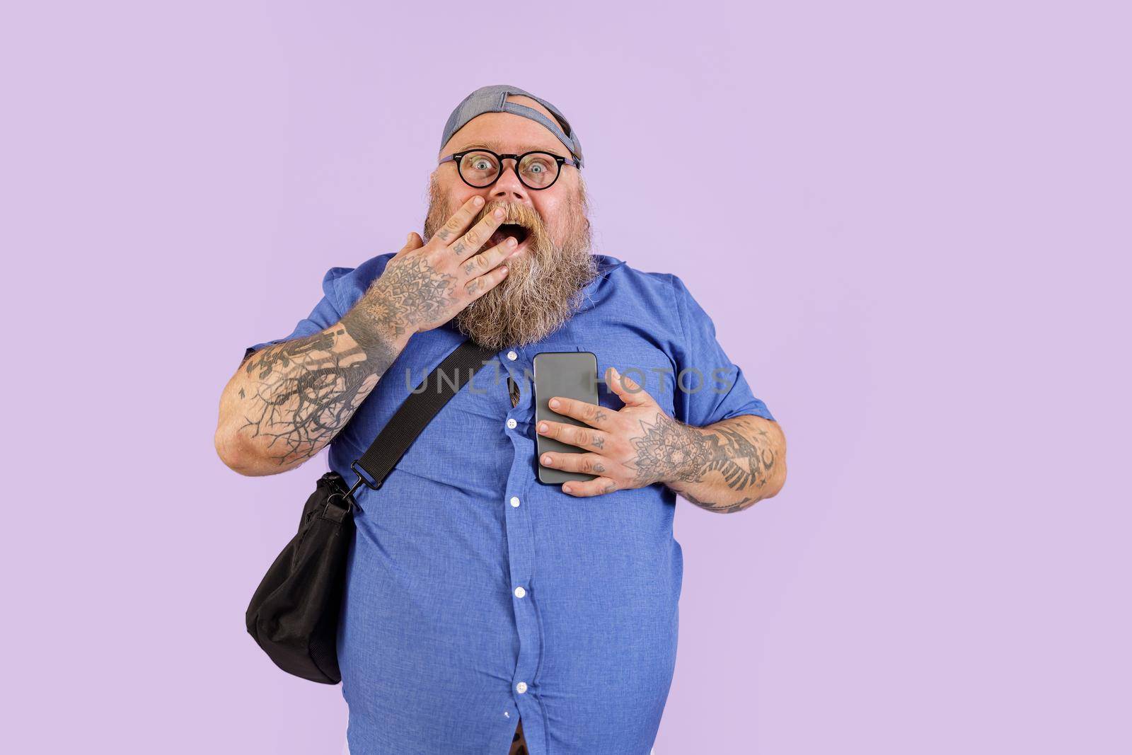 Emotional surprised obese man with crossbody bag and glasses holds mobile phone covering mouth by palm on purple background in studio
