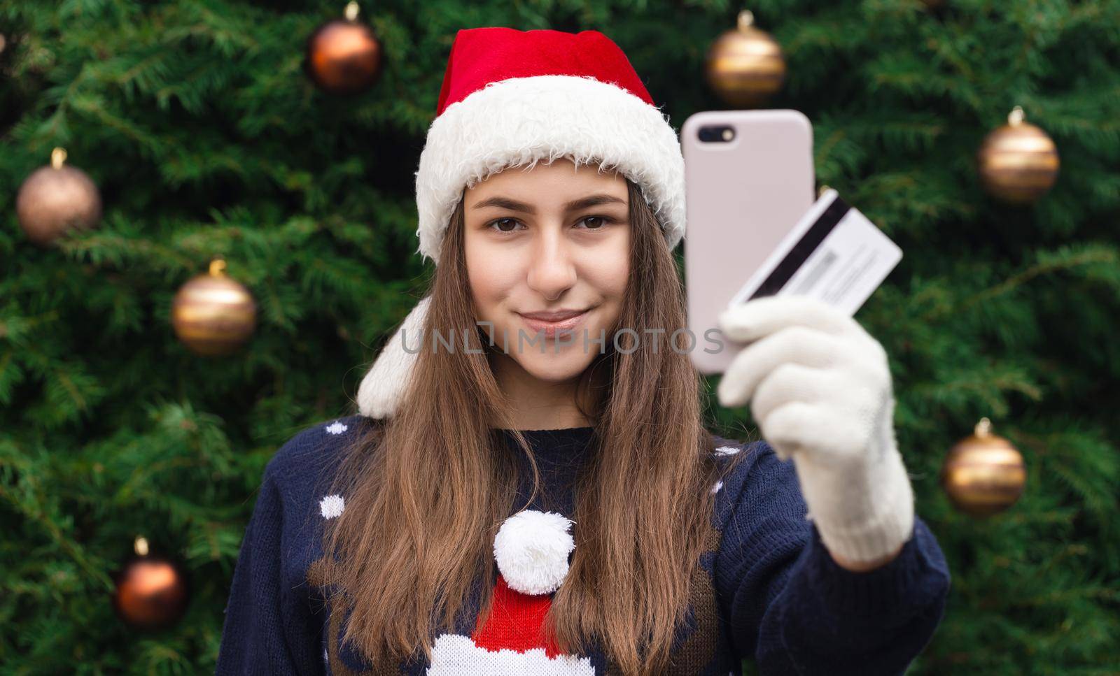 Christmas online shopping. A young girl in a santa claus hat and a blue sweater holding smartphone. Christmas during the coronavirus by lunarts