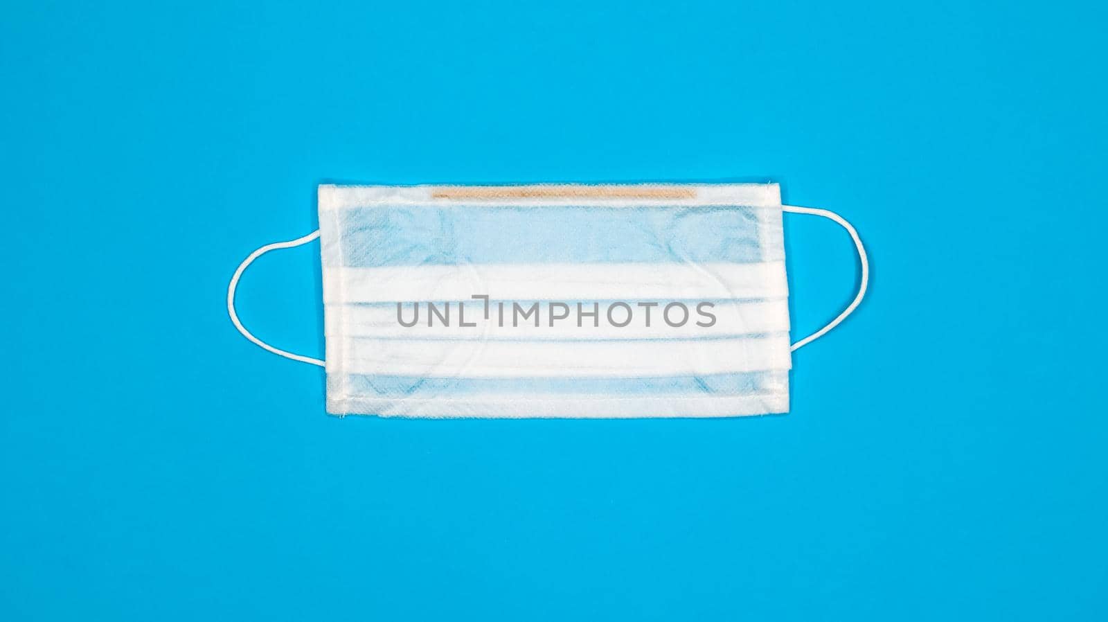 One 3-ply disposable surgical face mask with rubber grips covering mouth and nose against blue background. Protection from bacteria, healthcare and medicine concept. COVID-19. A dangerous virus