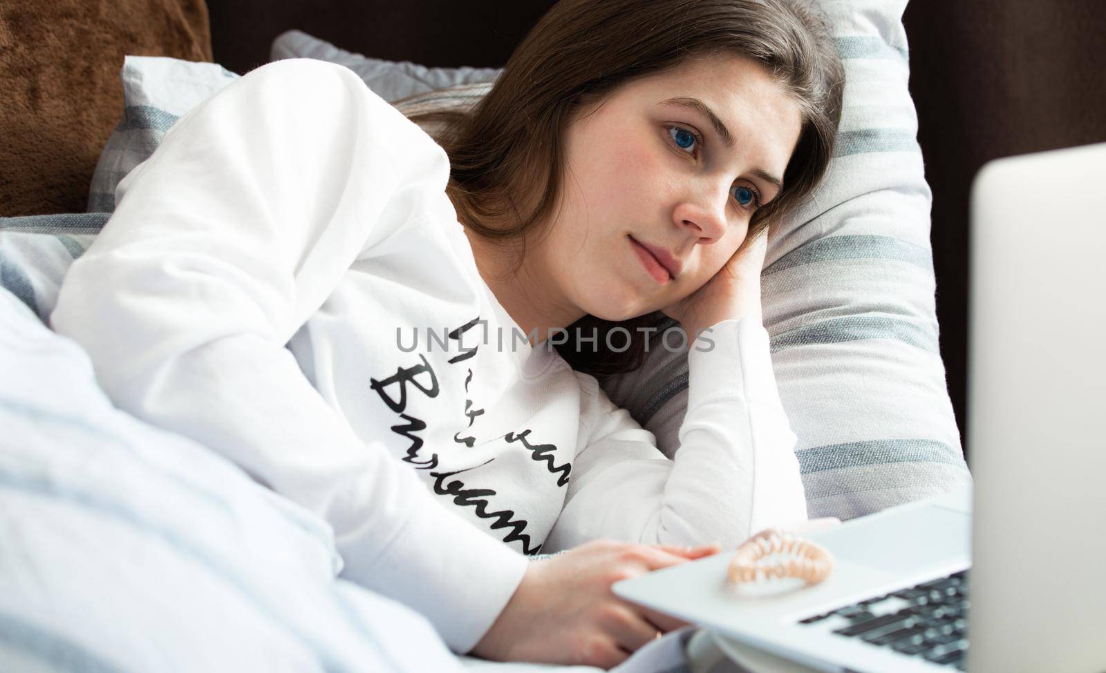 Young cute girl on a sunny day in a white jacket lies on the bed and looks into the laptop by lunarts