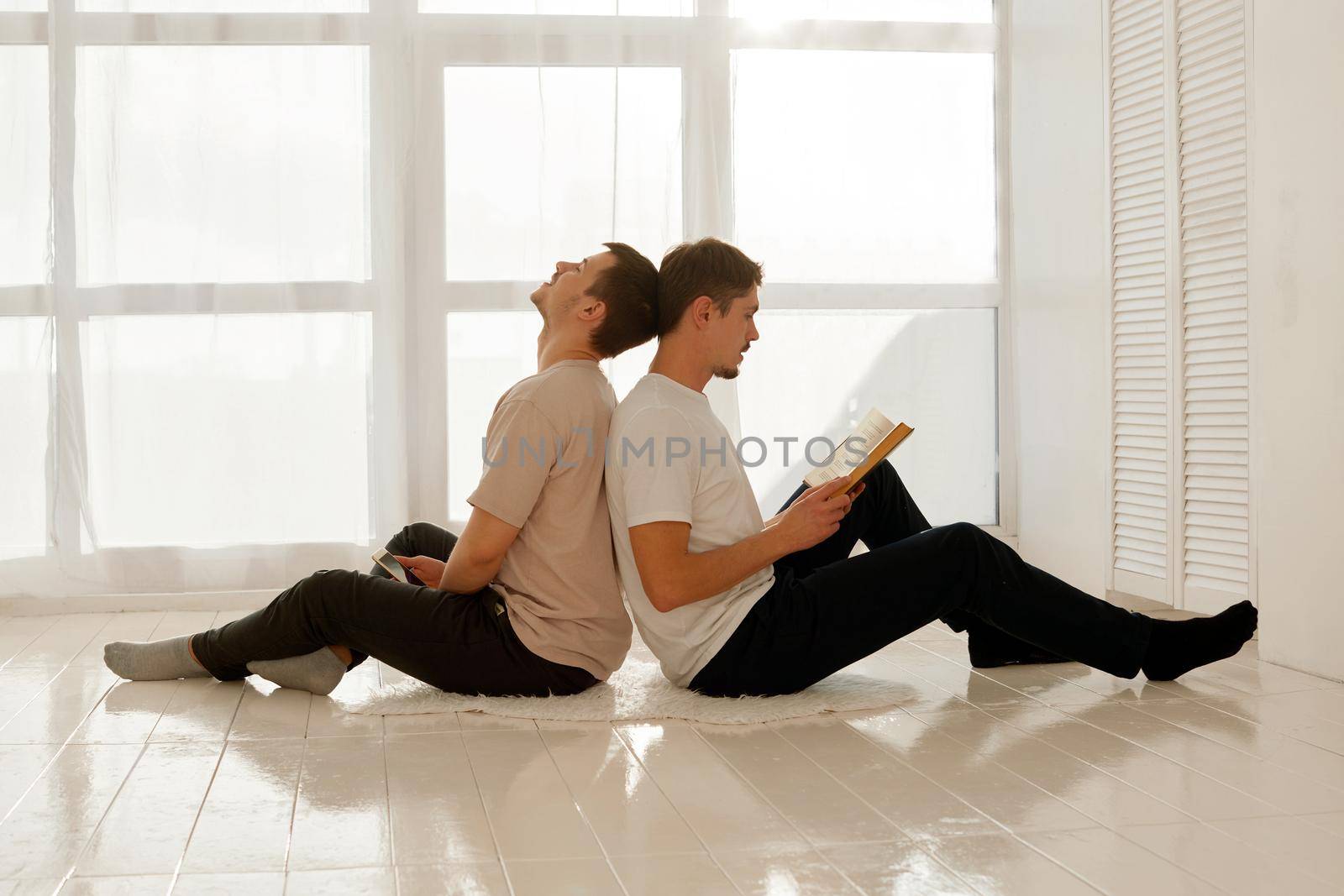 A nice guy reads a book and his friend listens to him and looks into a smartphone. Gay couple of men spend time at home sitting on the floor of the house in a light interior