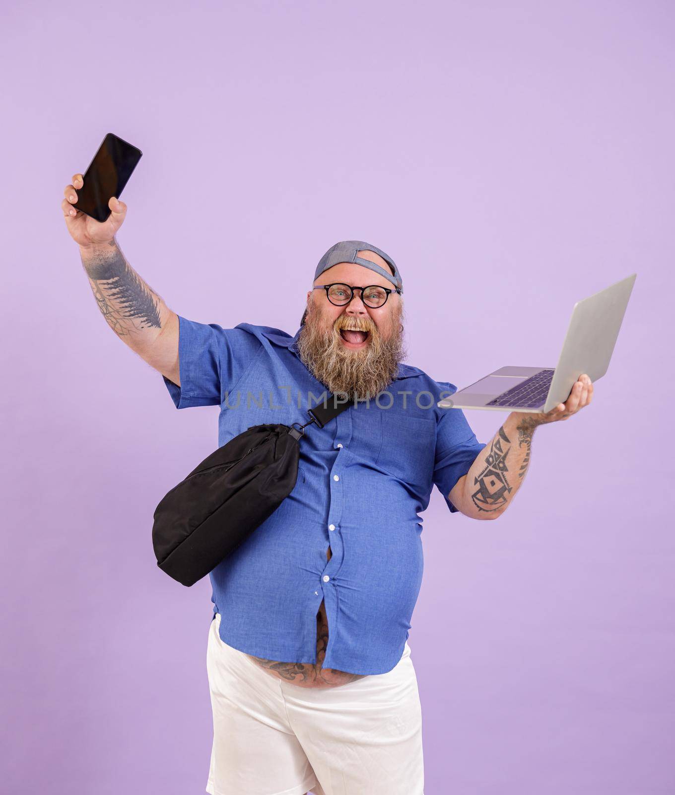 Positive plus size man with laptop and bag shows phone on purple background in studio by Yaroslav_astakhov