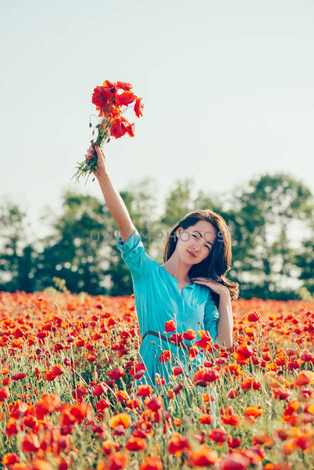 Beautiful young woman wearing in blue dress walking with bouquet in red poppy meadow.