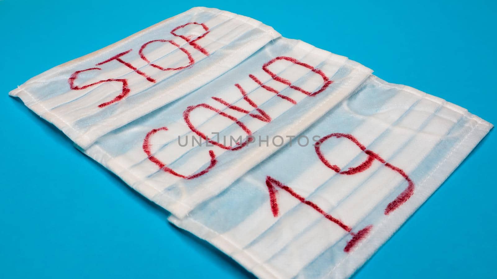 Typical 3-ply white surgical mask with rubber earhooks to cover mouth and nose with English block letters Stop Covid-19 on a blue background. Dangerous virus and quarantine concept.