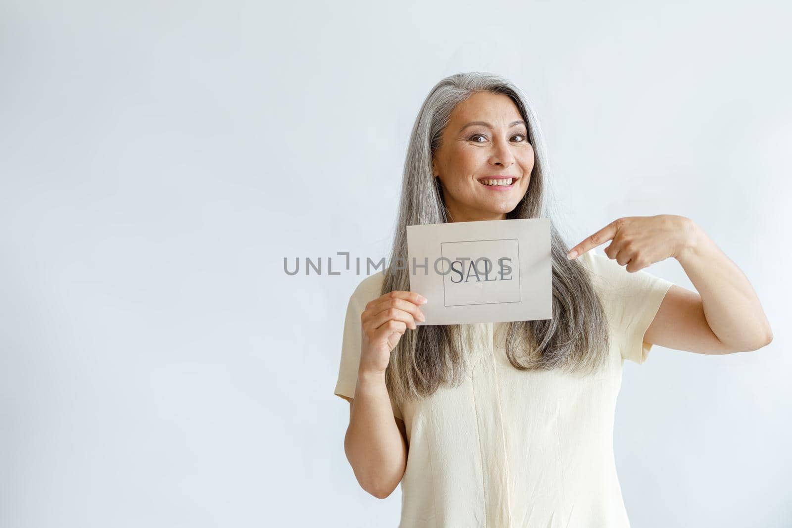 Joyful middle aged Asian woman with grey hair points onto Sale sign on light background by Yaroslav_astakhov