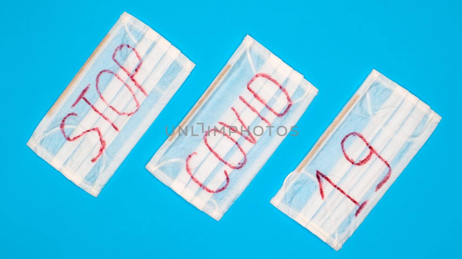 Typical 3-ply white surgical mask with rubber earhooks to cover mouth and nose with English block letters Stop Covid-19 on a blue background. Dangerous virus and quarantine concept.