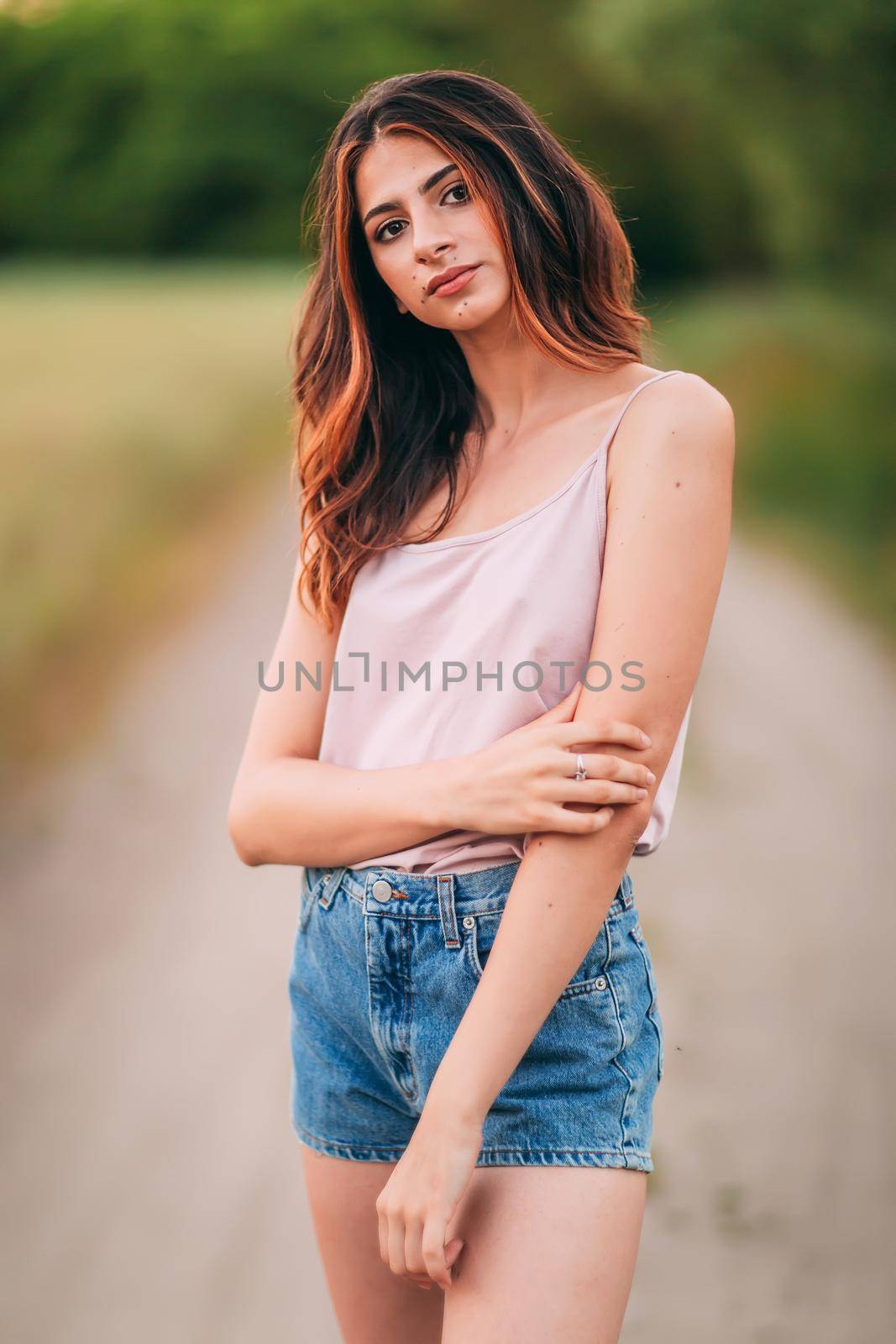 Portrait of beautiful sexy young woman with long brown hair in pink tank top and denim shorts posing outdoors at summer sunset, sensual, serious, blurry nature background