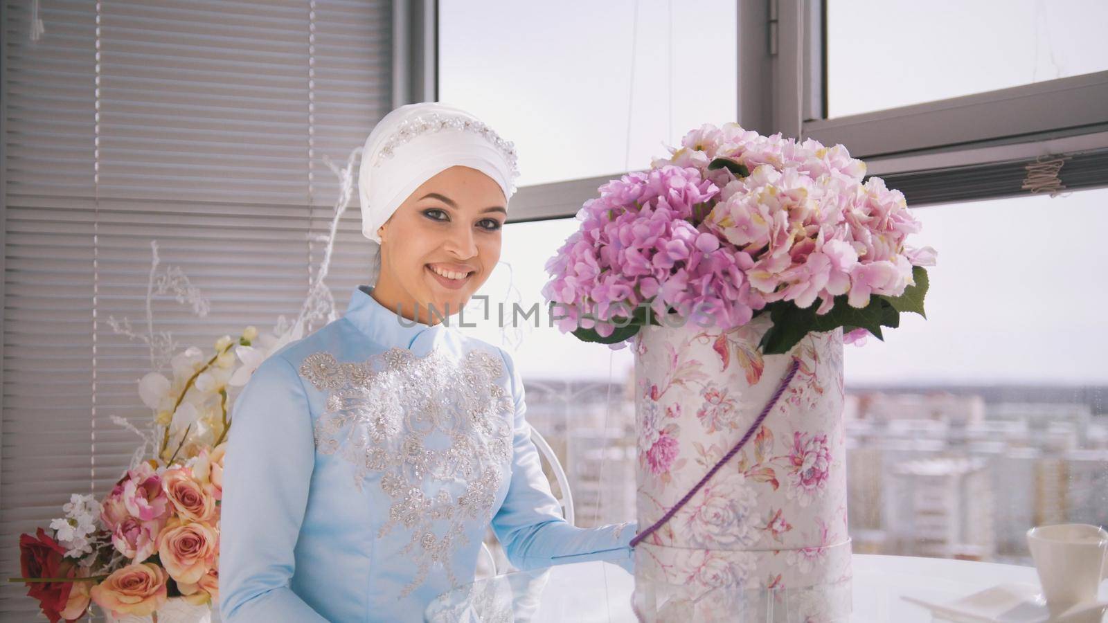 Portrait of smiling muslim bride in wedding dress with flowers sitting by the window, horizontal