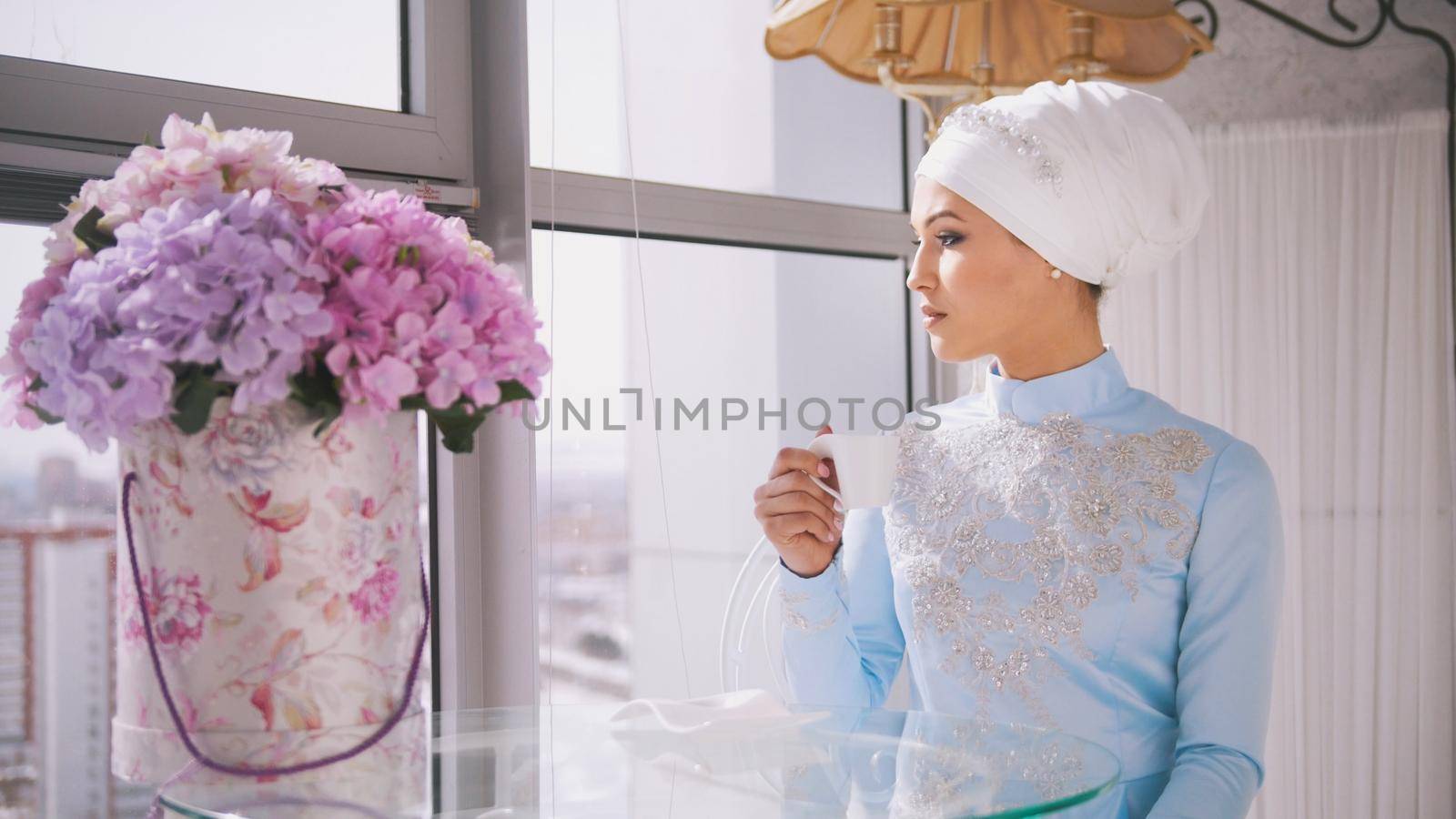 Beautiful bride in muslim wedding clothes with cup of coffee sitting by the window, horizontal