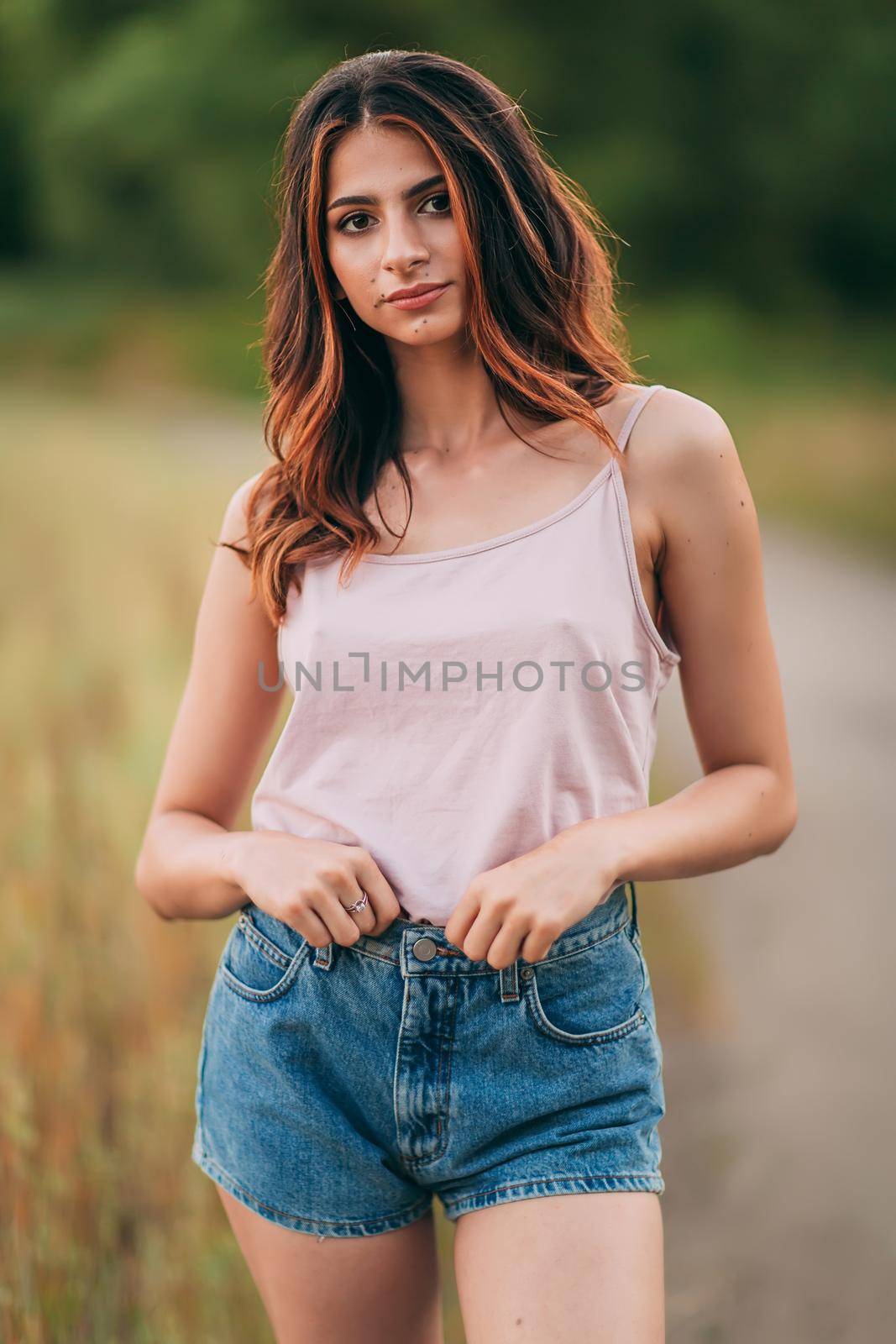 Portrait of beautiful sexy young woman with long brown hair in pink tank top and denim shorts posing outdoors at summer sunset, sensual, serious, blurry nature background