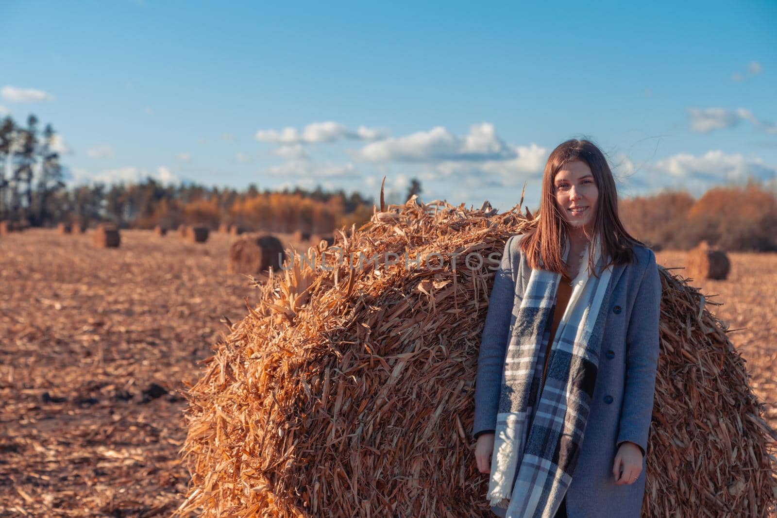 A girl of European appearance in a gray coat stands in a field near a larger bale with hay against a blue sky. by lunarts