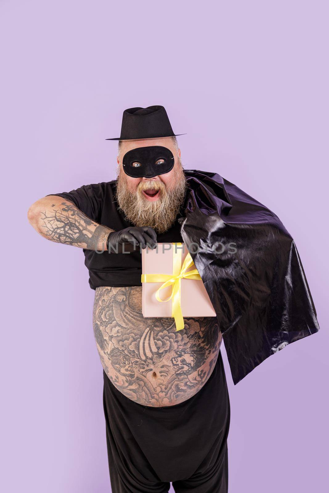 Excited man with overweight in Zorro costume hides gift box behind cape on purple background by Yaroslav_astakhov