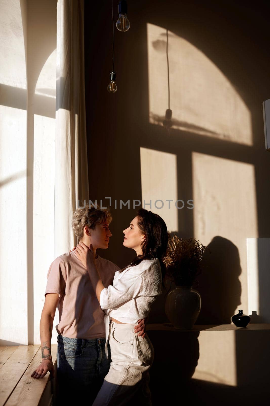 Side view of loving couple tenderly hugging while standing near windowsill in room lit by sunlight and looking at each other