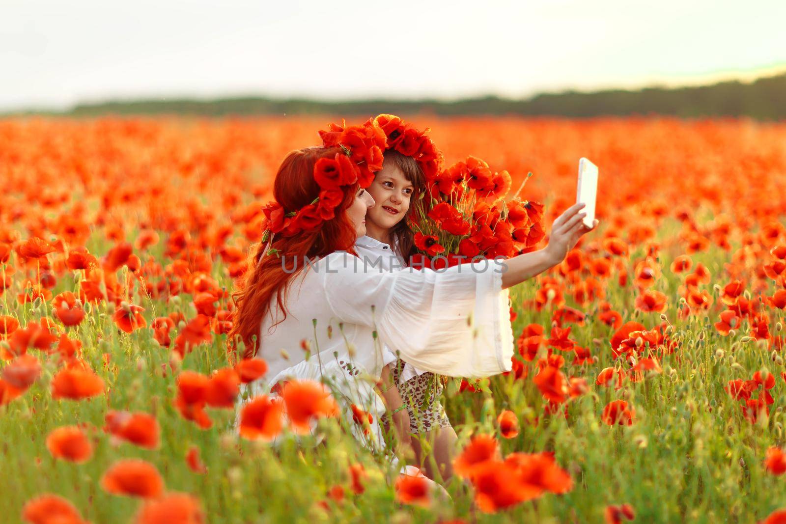 Little girl with redhead mother in white dresses and wreathes make selfie with bouquet of poppies on poppy field at warm summer sunset