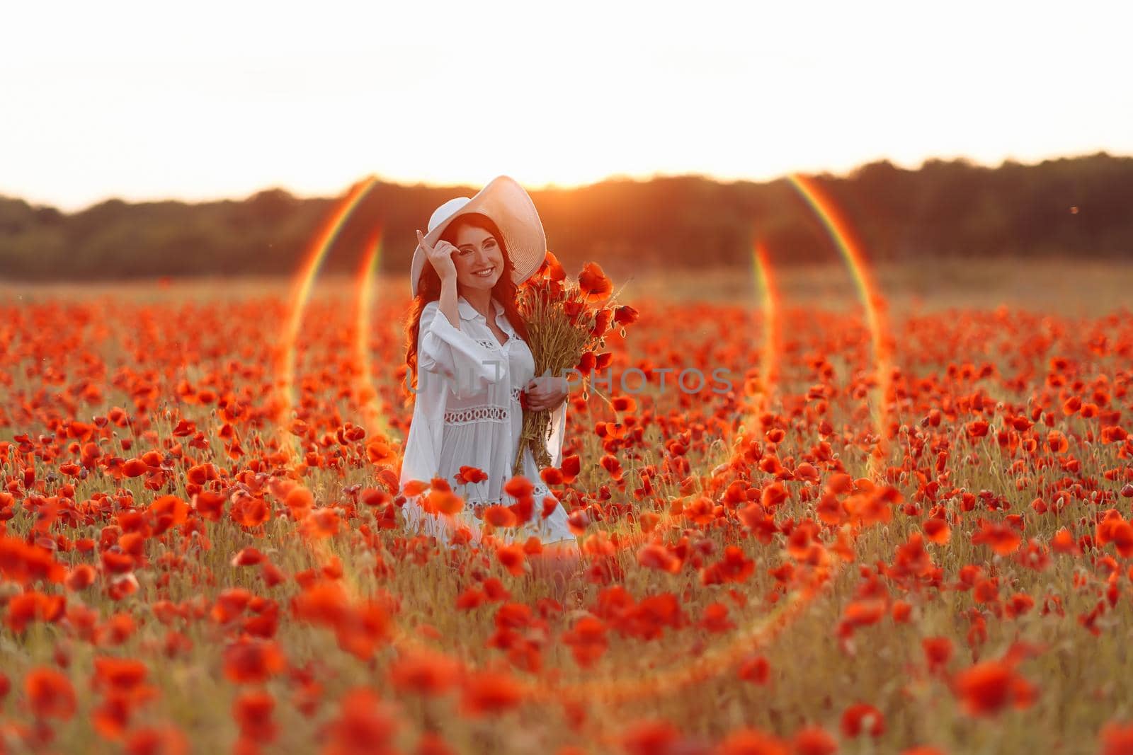Beautiful smiling happy redhead woman in white hat on green field with bouquet of poppies in summer sunset