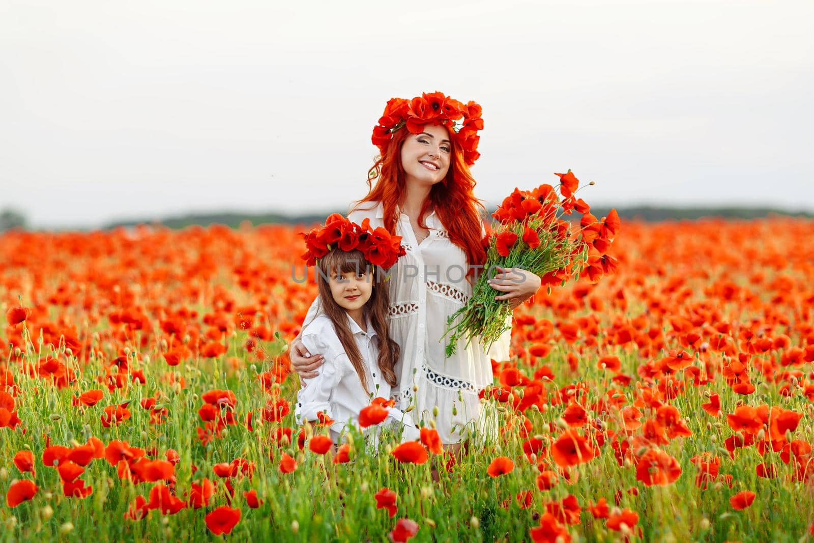 Little girl with redhead mother in white dresses and wreathes poses with bouquet of poppies on poppy field at warm summer sunset