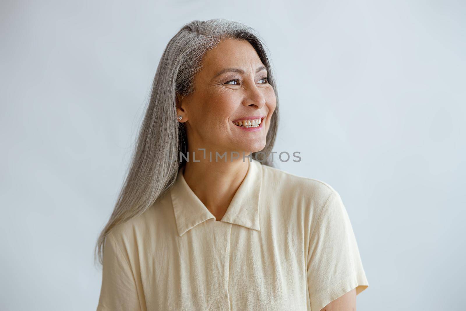 Joyful long haired middle aged lady wearing beige blouse stands on light background in studio. Mature beauty lifestyle