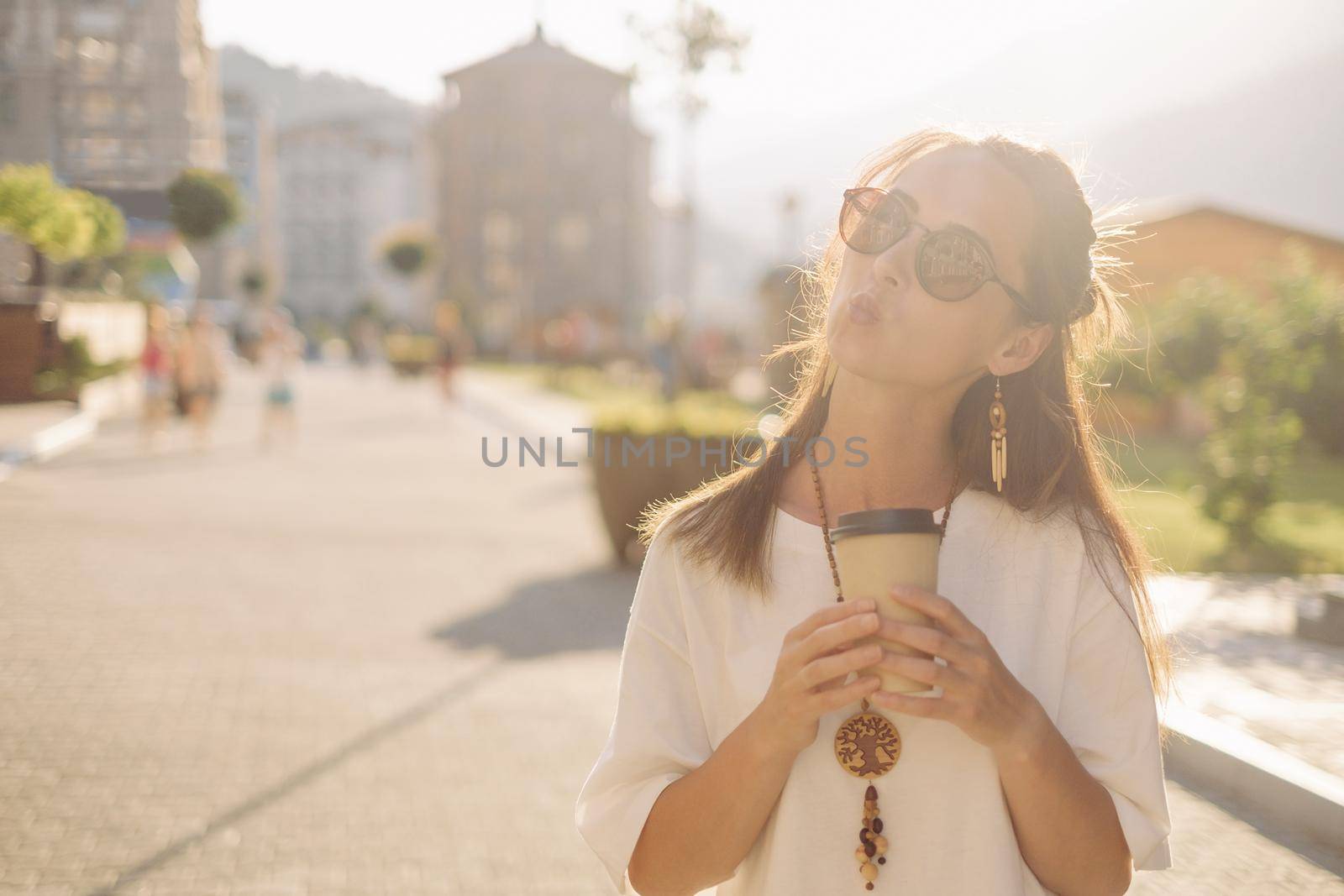 Beautiful young woman in white shirt and sunglasses walking with paper cup of coffee and making kiss gesture in the city.