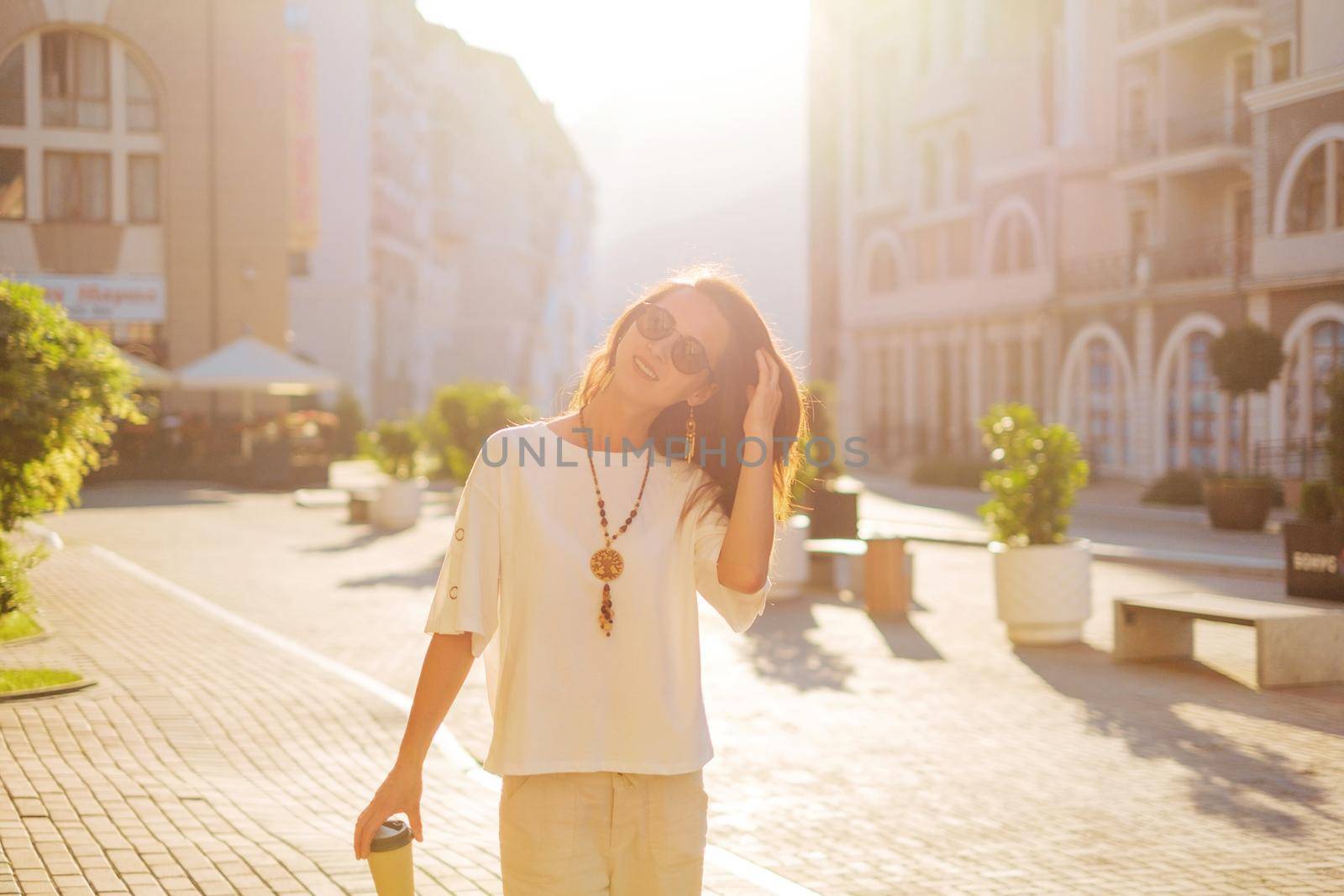 Cheerful stylish brunette young woman walking with paper cup of coffee in city street in spring morning.