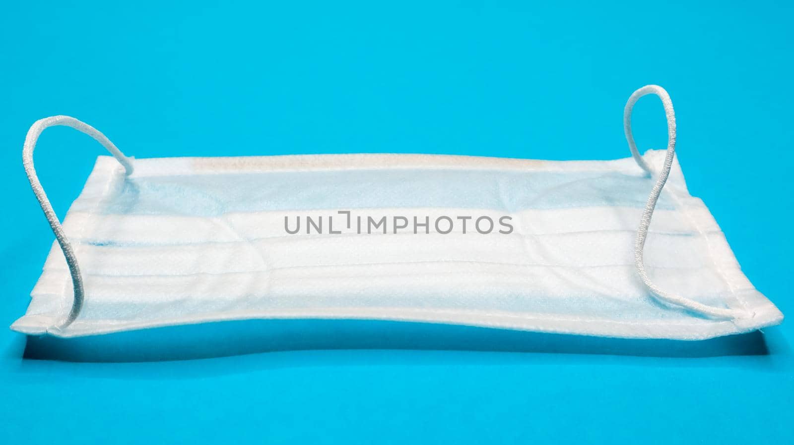 One 3-ply disposable surgical face mask with rubber grips covering mouth and nose against blue background. Protection from bacteria, healthcare and medicine concept. COVID-19. A dangerous virus