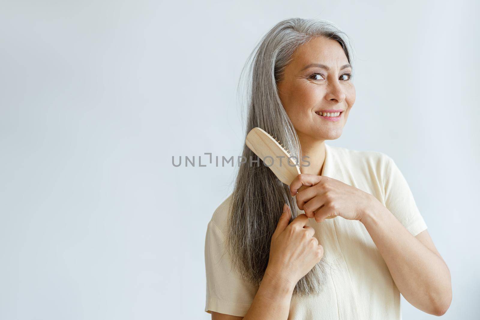 Thoughtful middle aged Asian woman brushes grey hair posing on light background by Yaroslav_astakhov