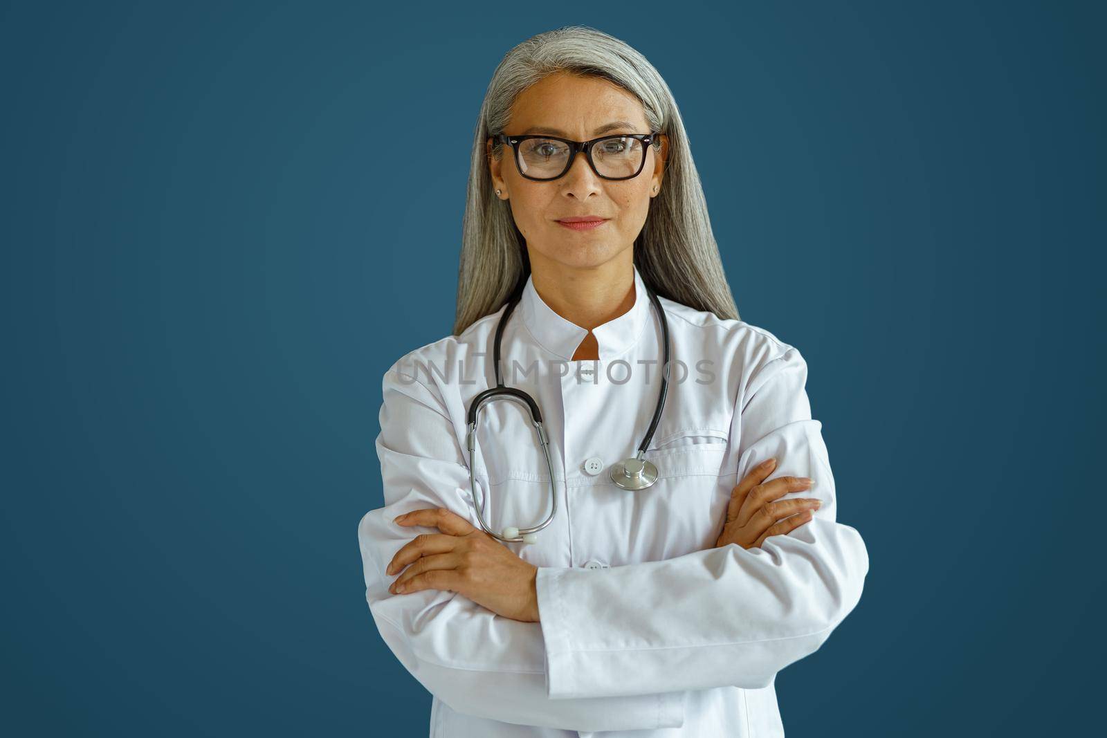 Mature female doctor in white robe with glasses and stethoscope stands on blue background in studio. Professional medical staff