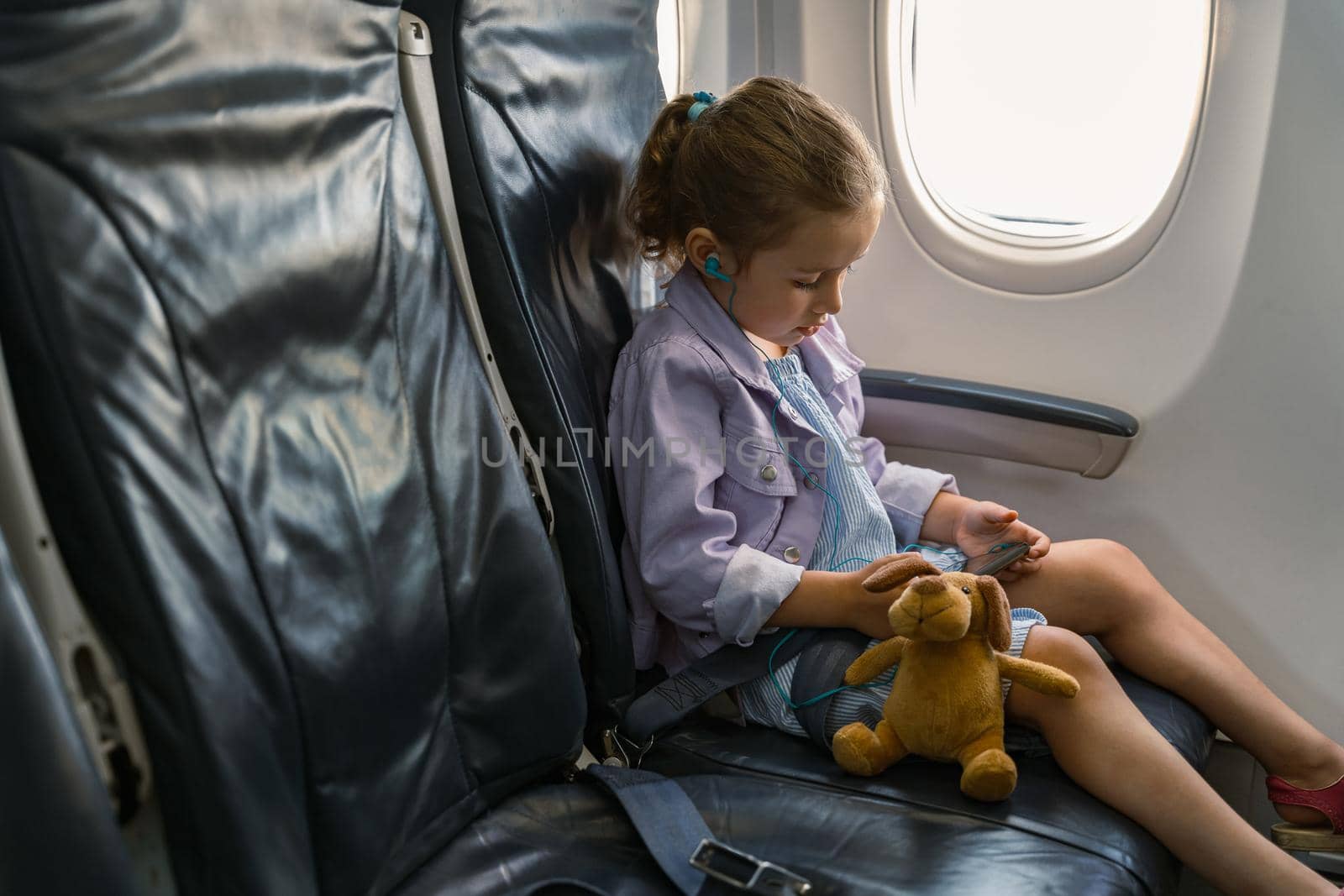 Little girl sitting in a chair with toy and holding a phone during trip by plane by Yaroslav_astakhov