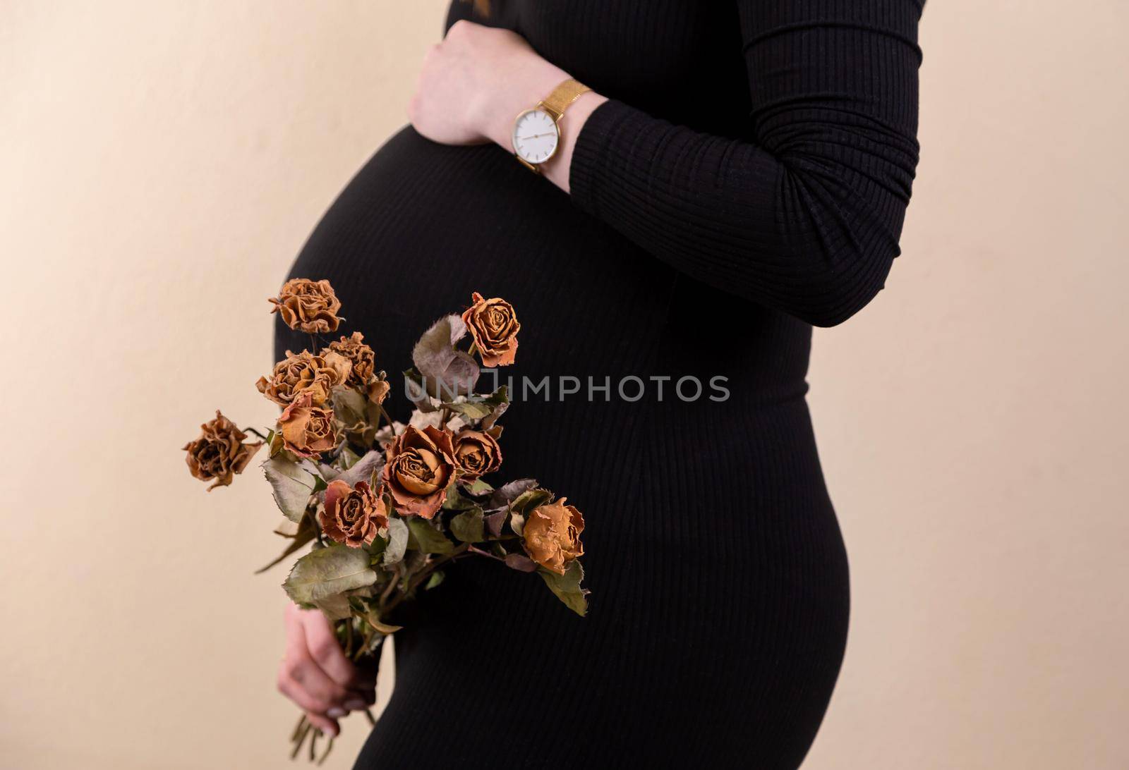 Side view of an attractive pregnant woman caressing her belly and holding a bouquet of dry roses in black dress while standing against yellow background by lunarts