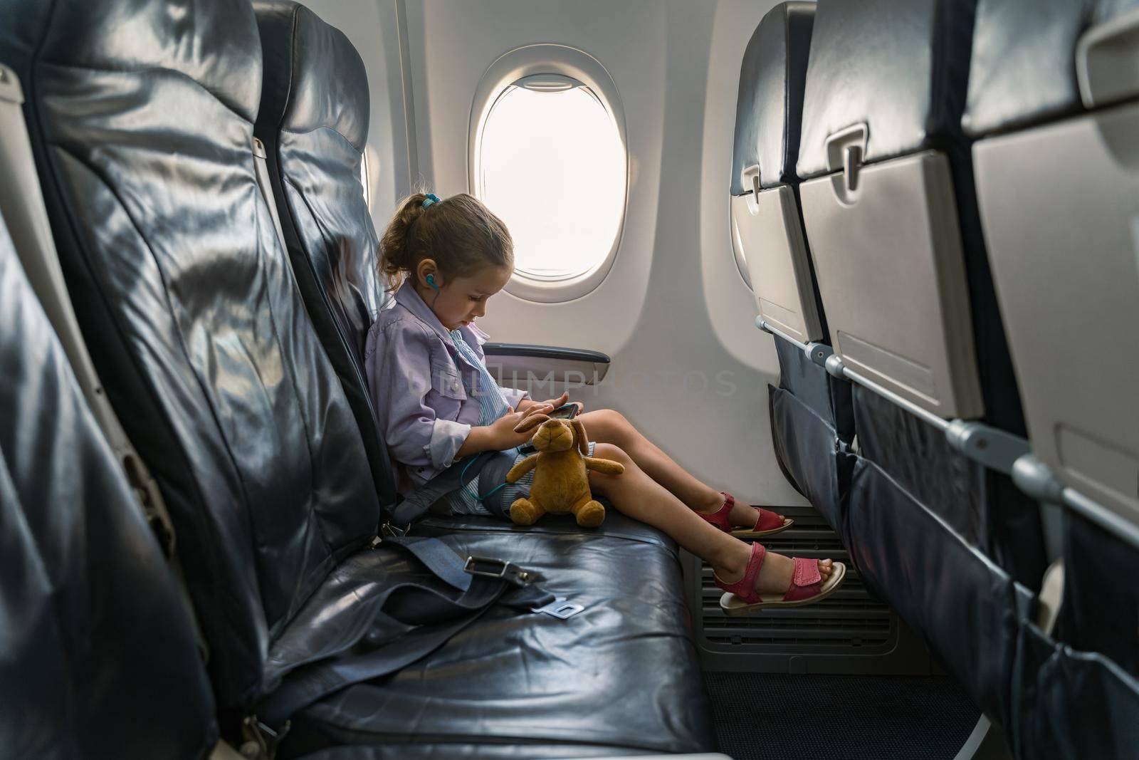 Side view of cute kid looking at mobile phone while using headphones in plane. Trip concept