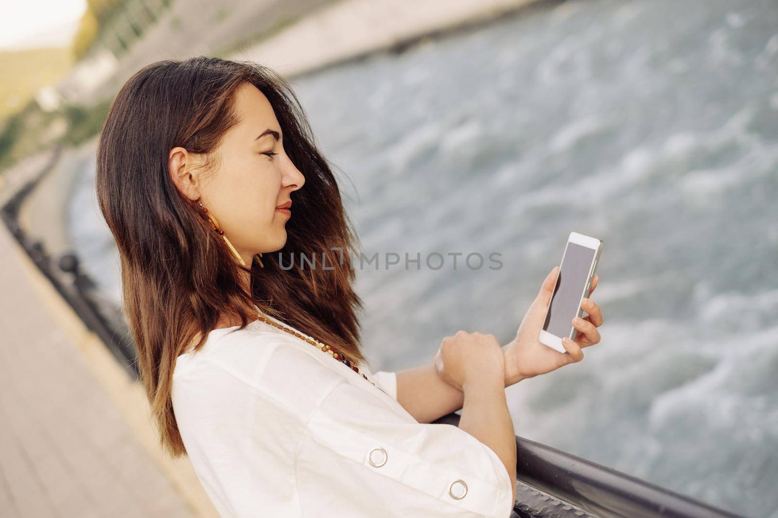 Smiling young woman using mobile phone while standing on embankment of river in summer outdoor.