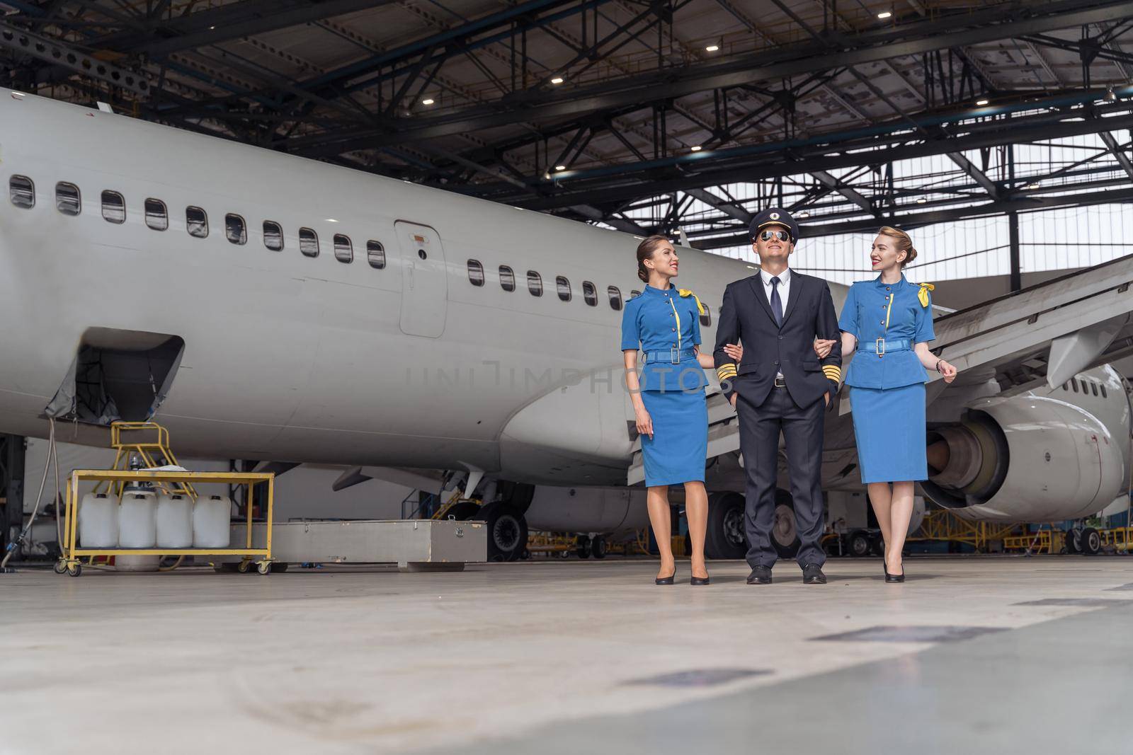 Happy pilot with two air stewardesses walking near airplane in hangar