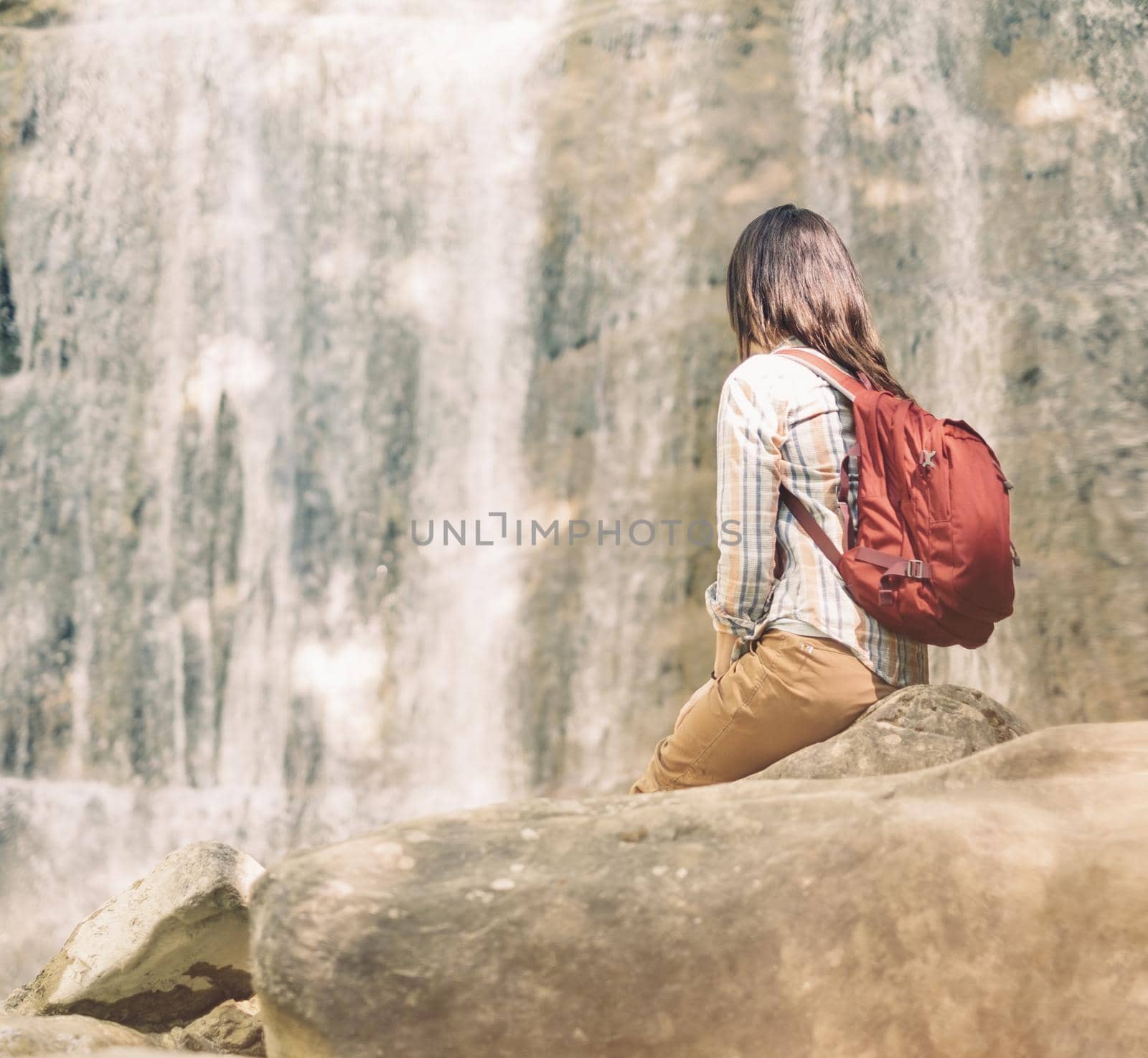 Unrecognizable traveler backpacker young woman sitting on stone and looking at waterfall on sunny day outdoor.