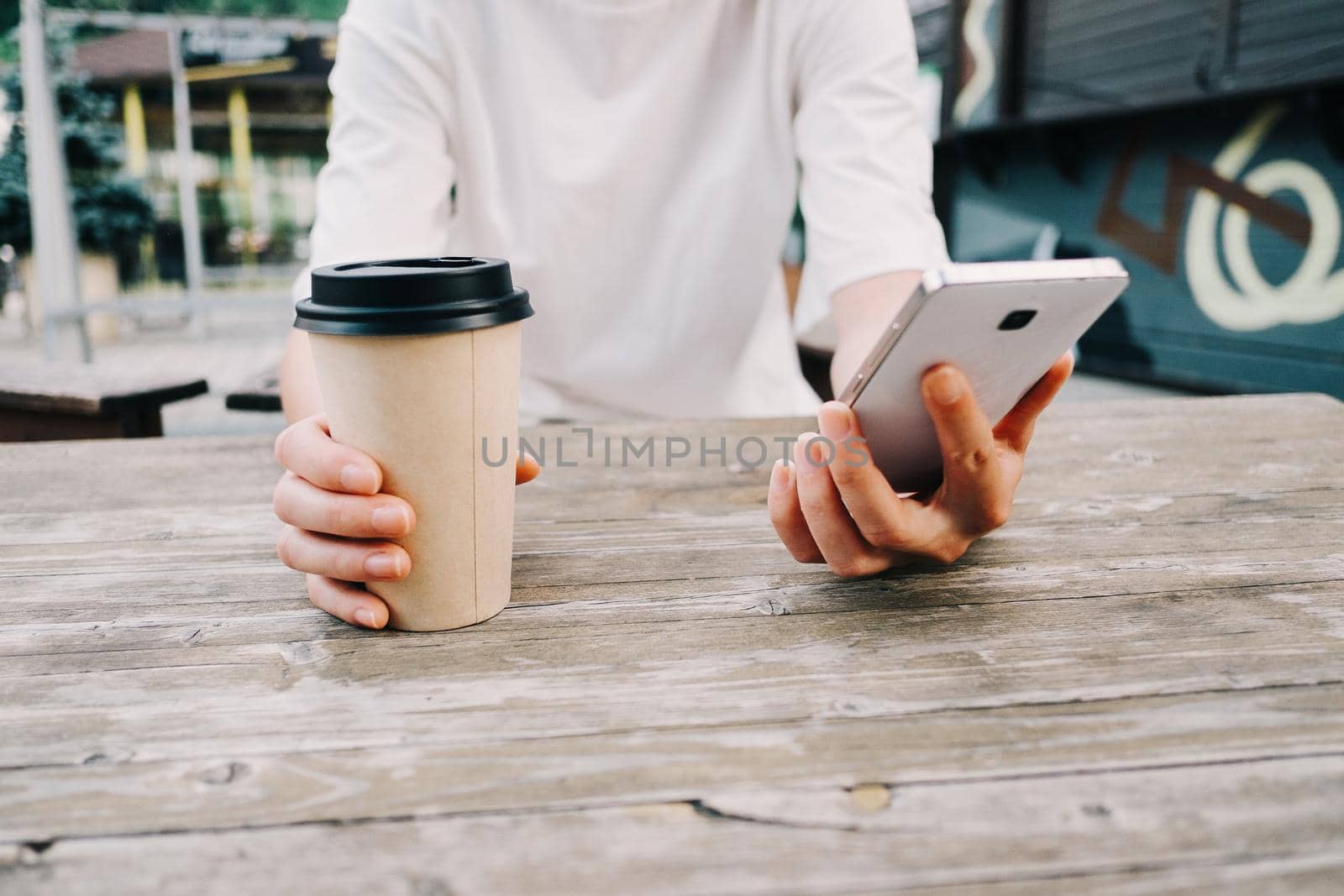 Unrecognizable young woman sitting with cup of coffee and mobile phone at wooden table in cafe in city street.