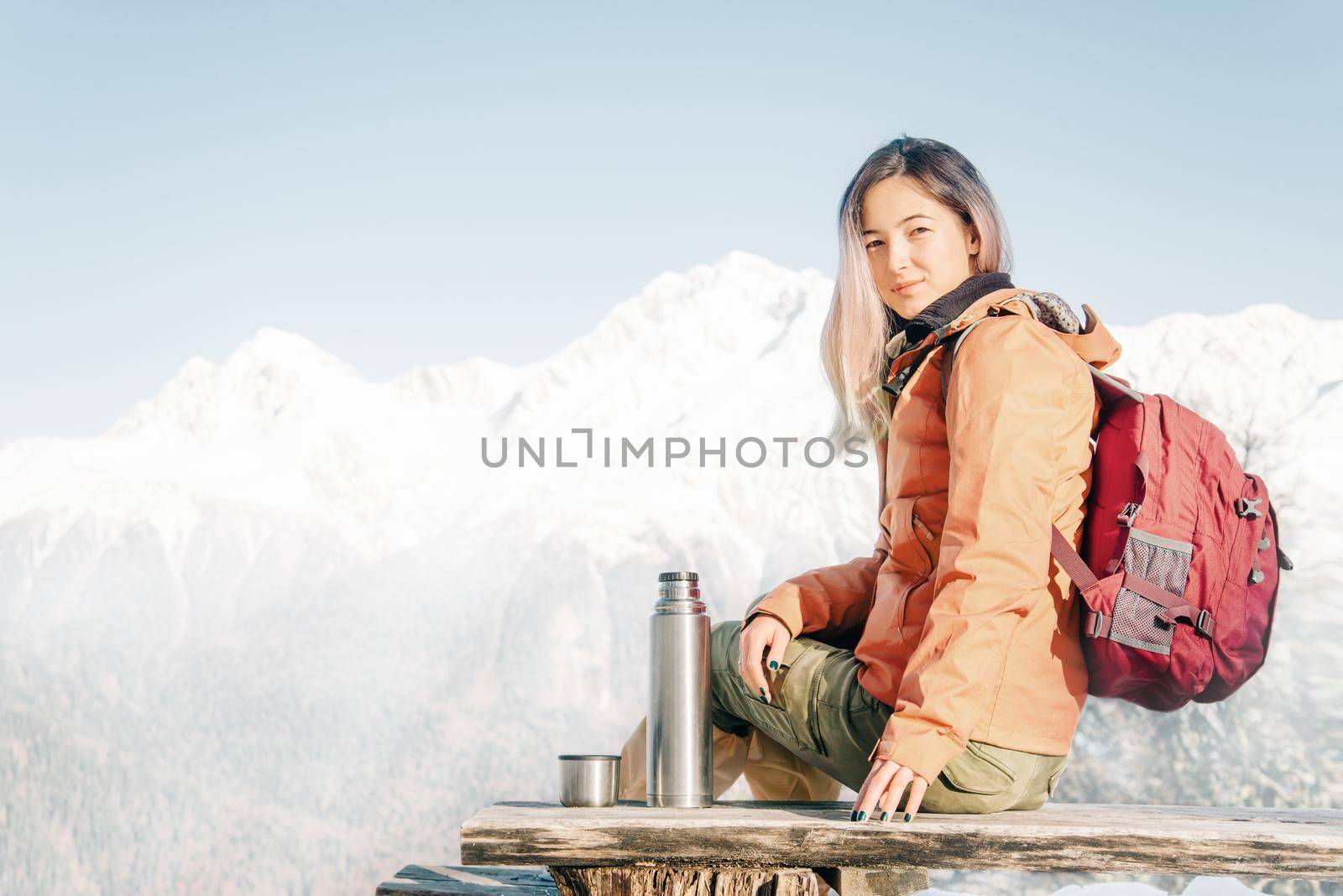 Backpacker explorer young woman sitting on wooden bench near the thermos on background of mountains in winter.