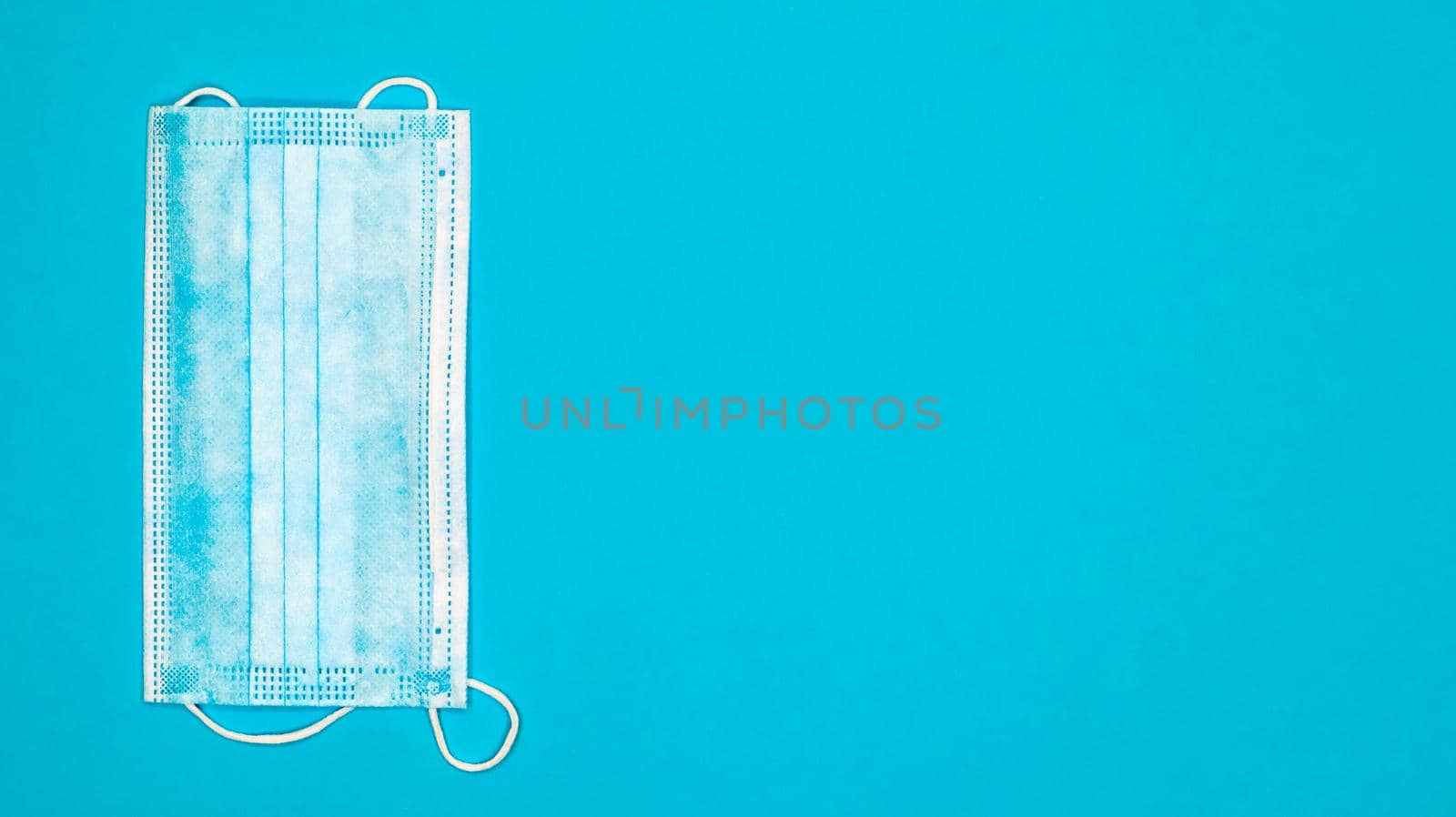 One 3-ply disposable surgical face mask with rubber pads covering the mouth and nose against a blue background. The concept of protection from bacteria, healthcare and medicine. COVID-19. copy space.