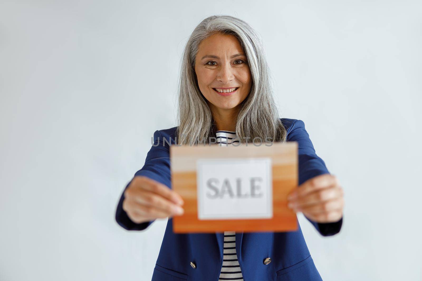 Attractive middle aged Asian woman with natural grey hair in stylish jacket holds card with word Sale standing on light background in studio