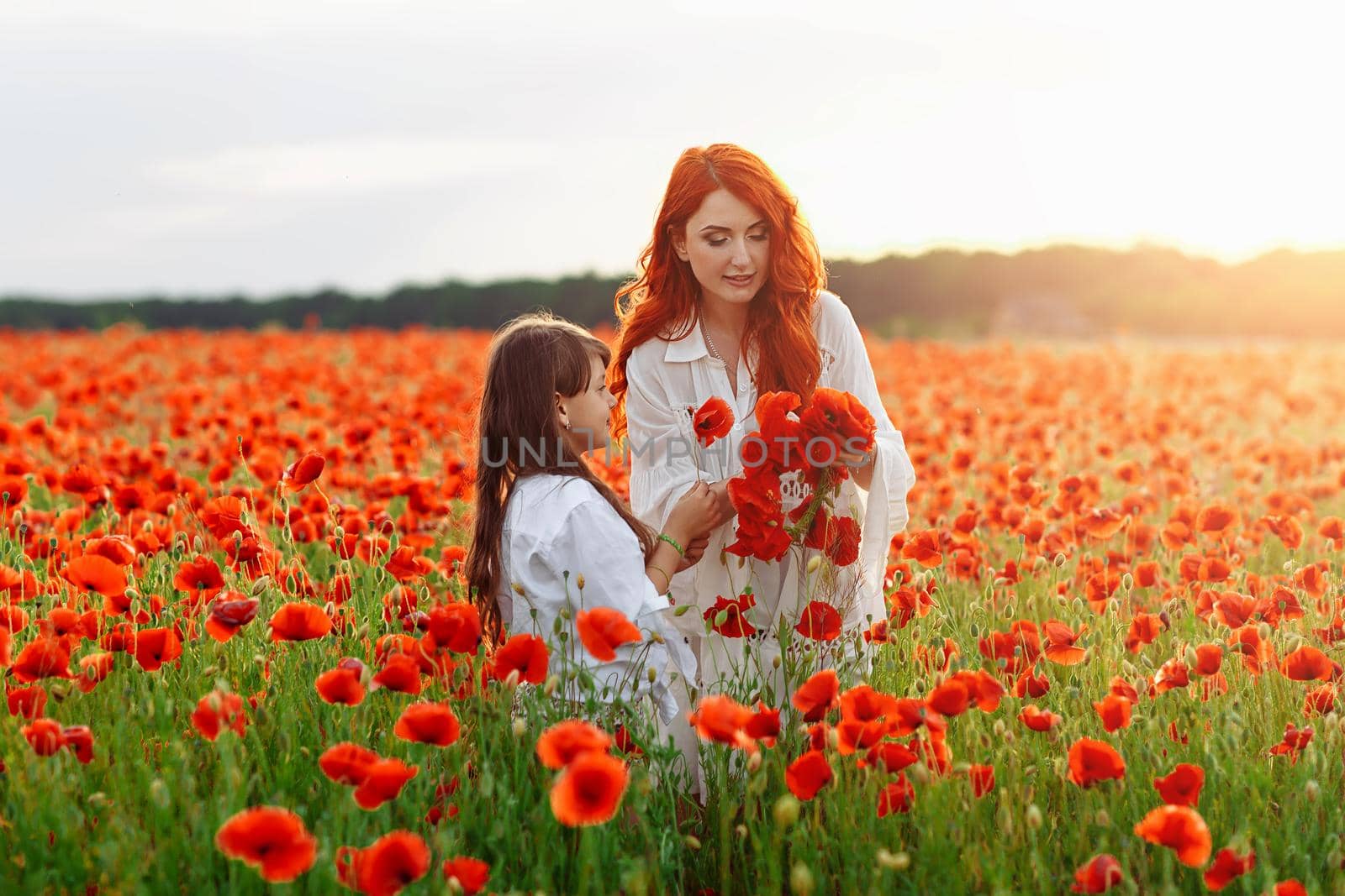 Little happy girl with redhead mother in white dresses makes wreath on poppy field at warm summer sunset