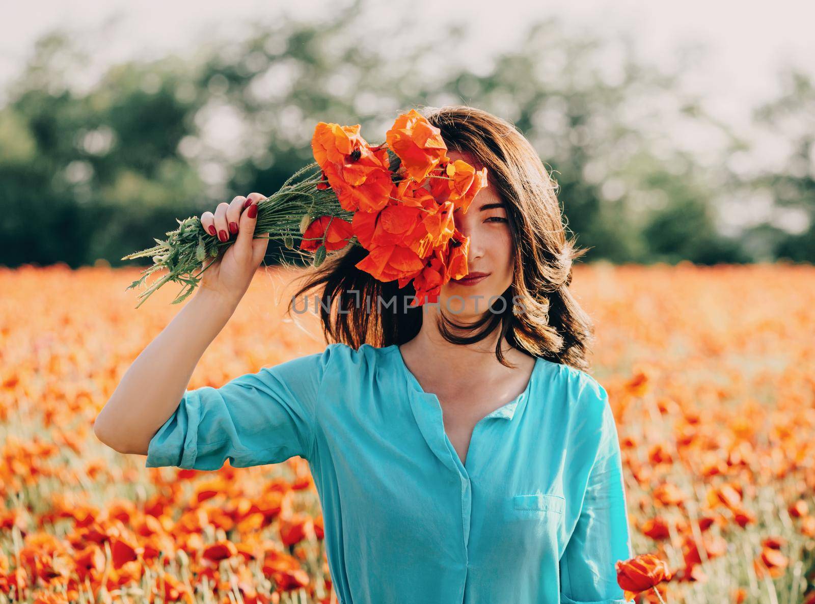 Beautiful young woman standing with red poppies bouquet in flower meadow in spring outdoor, looking at camera.