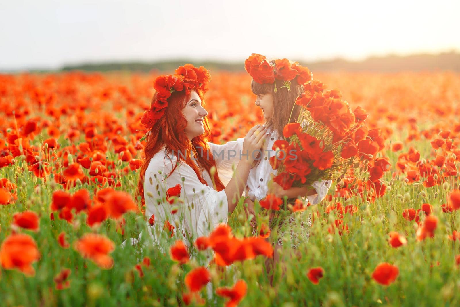 Little girl with redhead mother in white dresses and wreathes poses with bouquet of poppies on poppy field at warm summer sunset