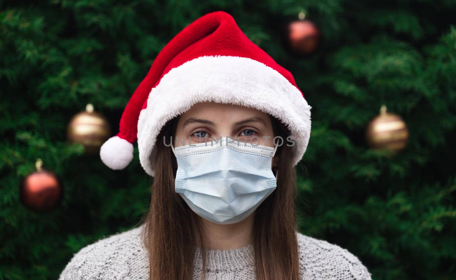 Close up Portrait of woman wearing a santa claus hat and medical mask with emotion. Against the background of a Christmas tree. Coronavirus pandemic by lunarts